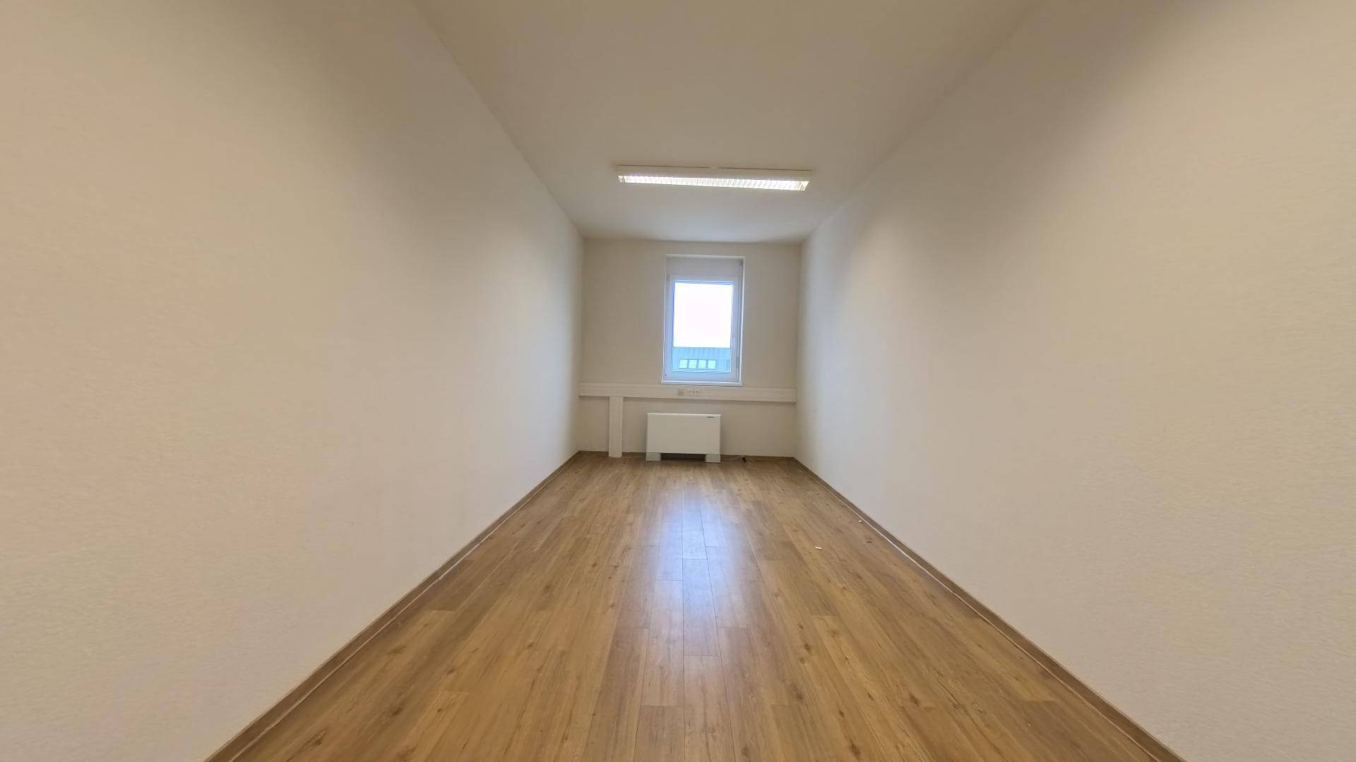 Langer, leerer Raum mit hellem Laminatboden, weißen Wänden und einem Fenster am Ende, das Tageslicht hereinlässt.
