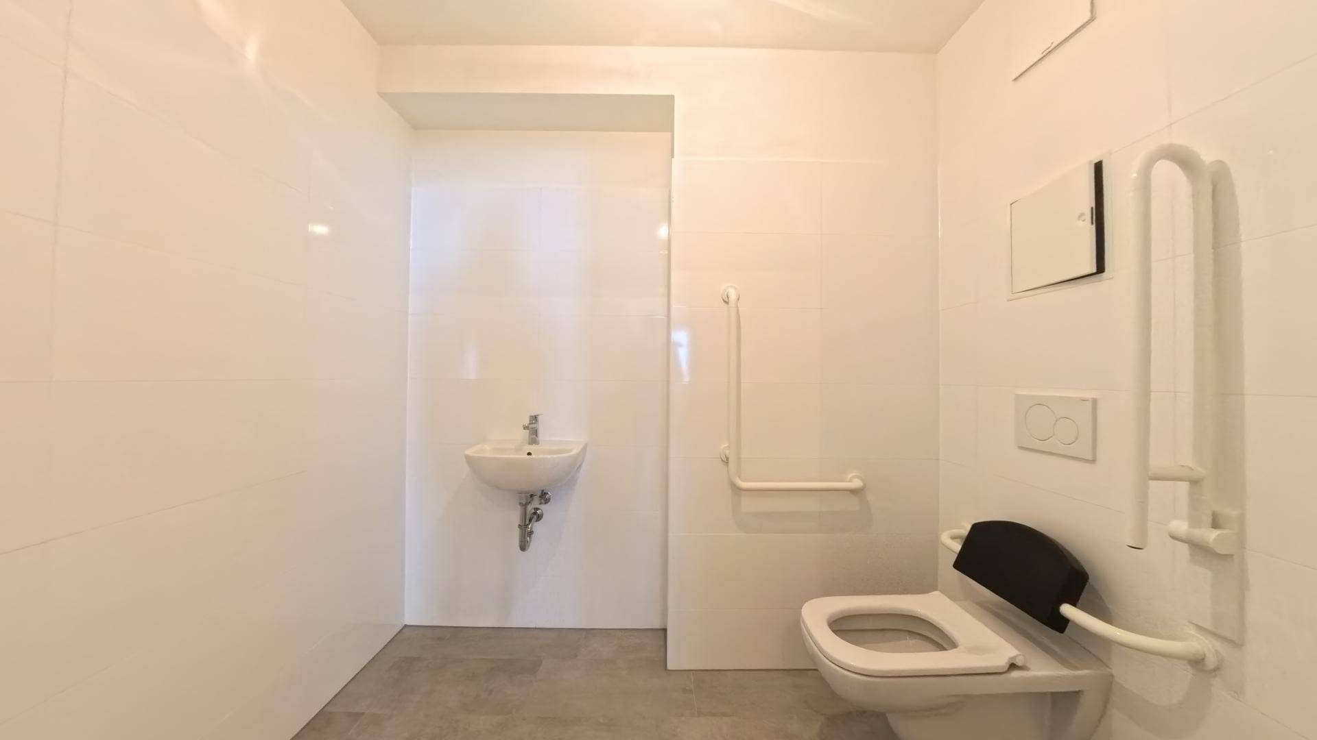 Modernes WC mit weißen Fliesen, kleinem Waschbecken und Haltegriffen für Barrierefreiheit.