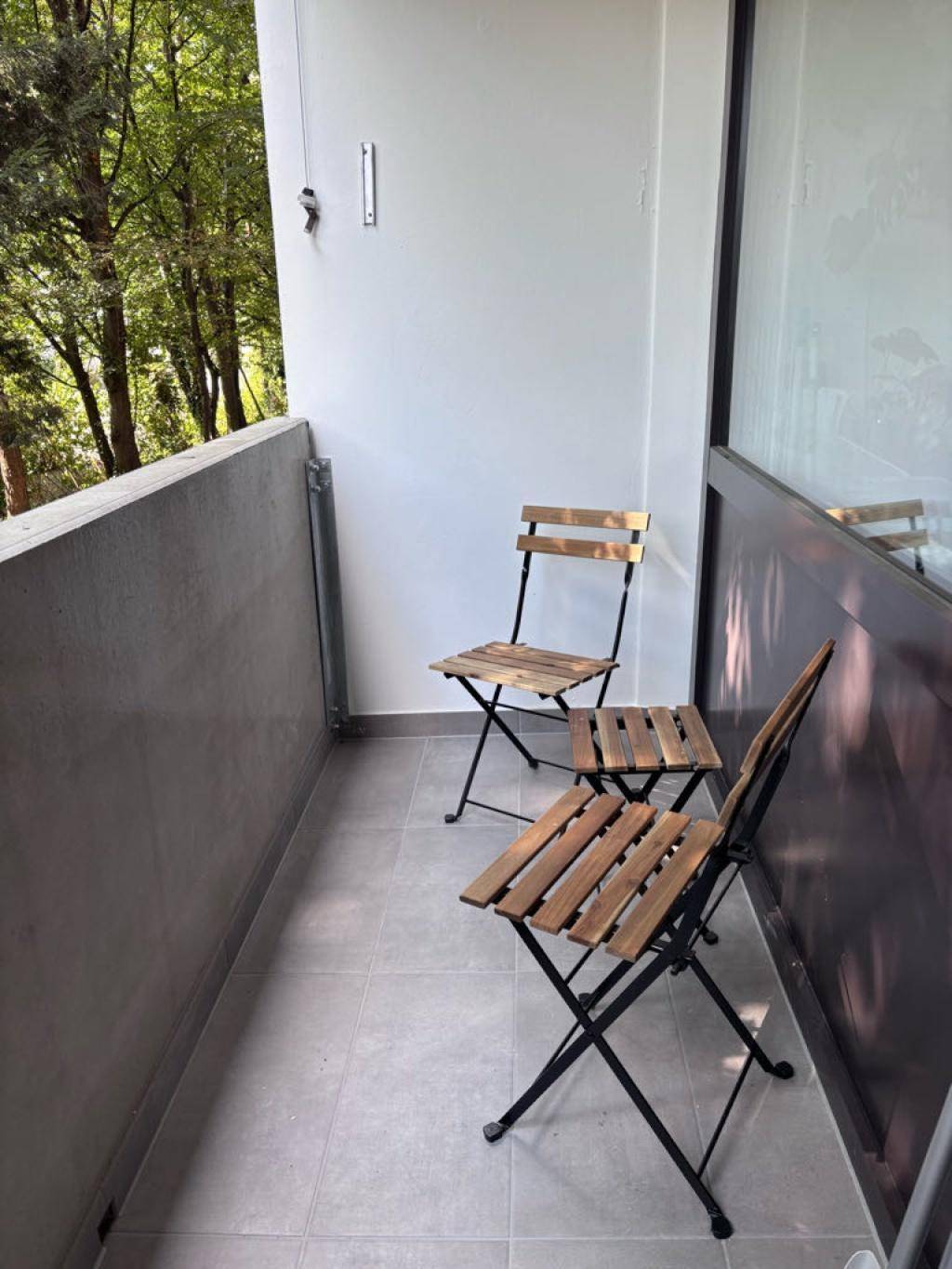Schmaler Balkon mit Fliesenboden und zwei Holzstühlen, umgeben von Bäumen.