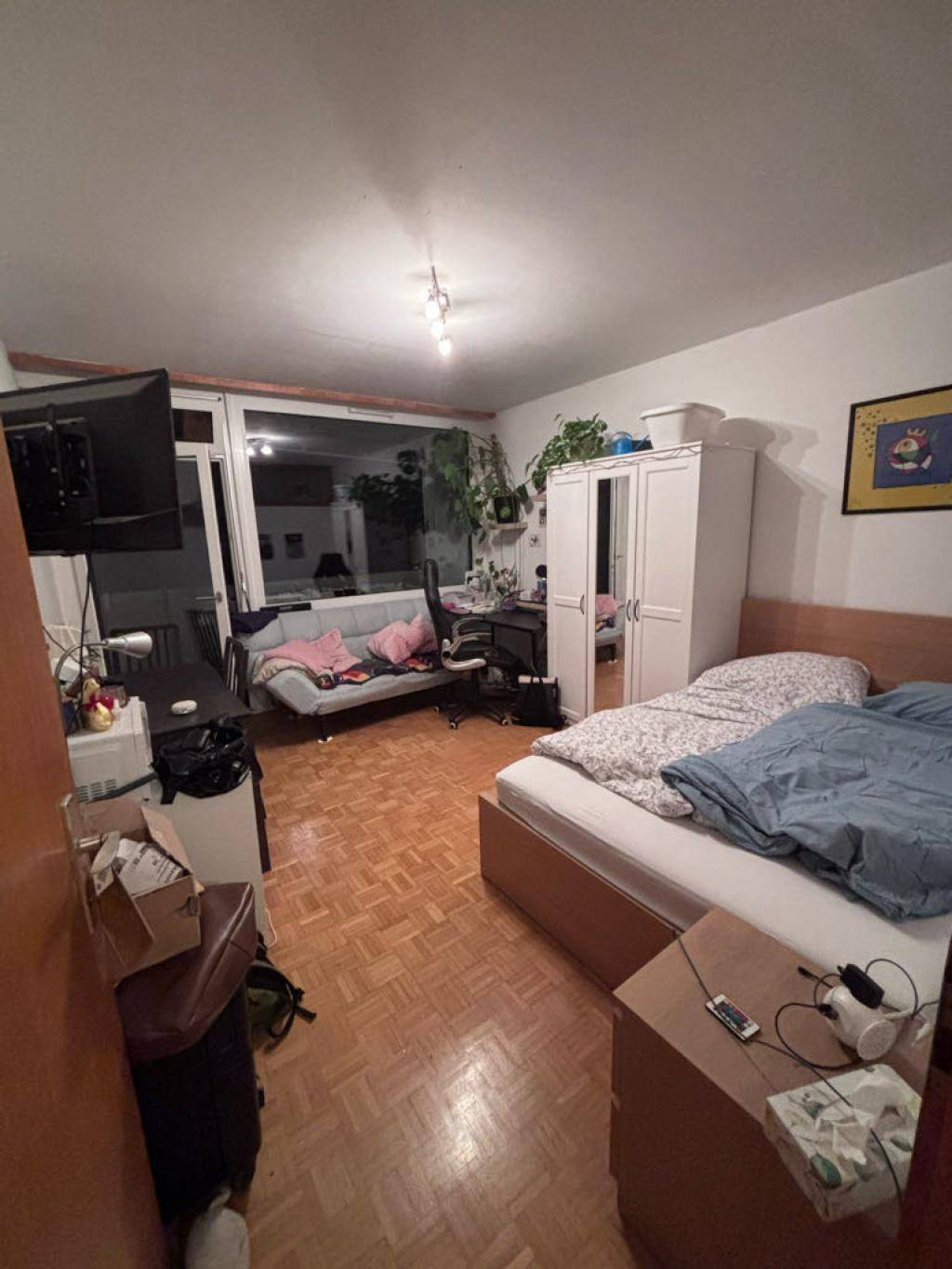 Schlafzimmer mit Bett, Schreibtisch und Zugang zu einem Balkon, abends beleuchtet.