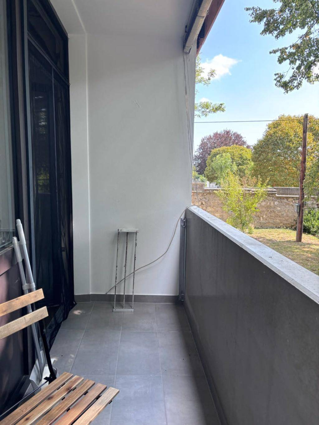 Balkon mit Fliesenboden und Blick auf Bäume und eine Mauer im Hintergrund.