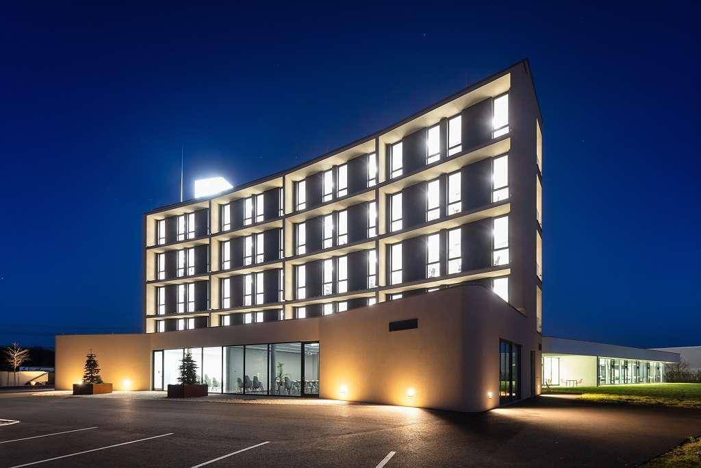 Modernes Gebäude bei Nacht, hell beleuchtet mit vielen Fenstern und einem großen Parkplatz.