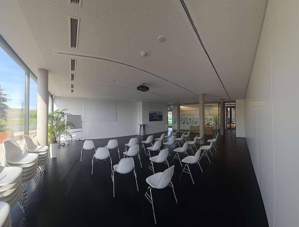 Geräumiger Konferenzraum mit vielen weißen Stühlen, großen Fenstern und einer Projektionsfläche.