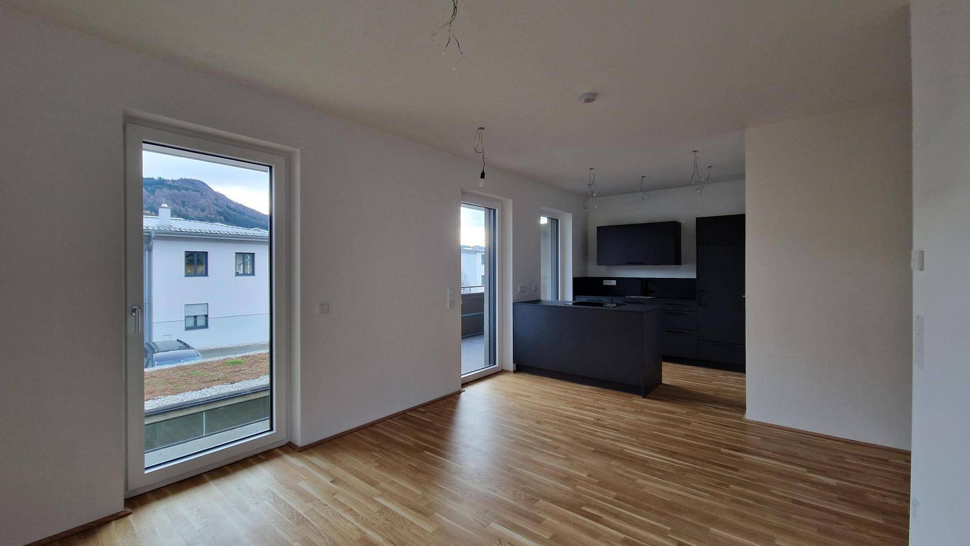 Heller offener Wohn-Essbereich mit Holzboden und großen Fenstern, die Zugang zum Balkon bieten.