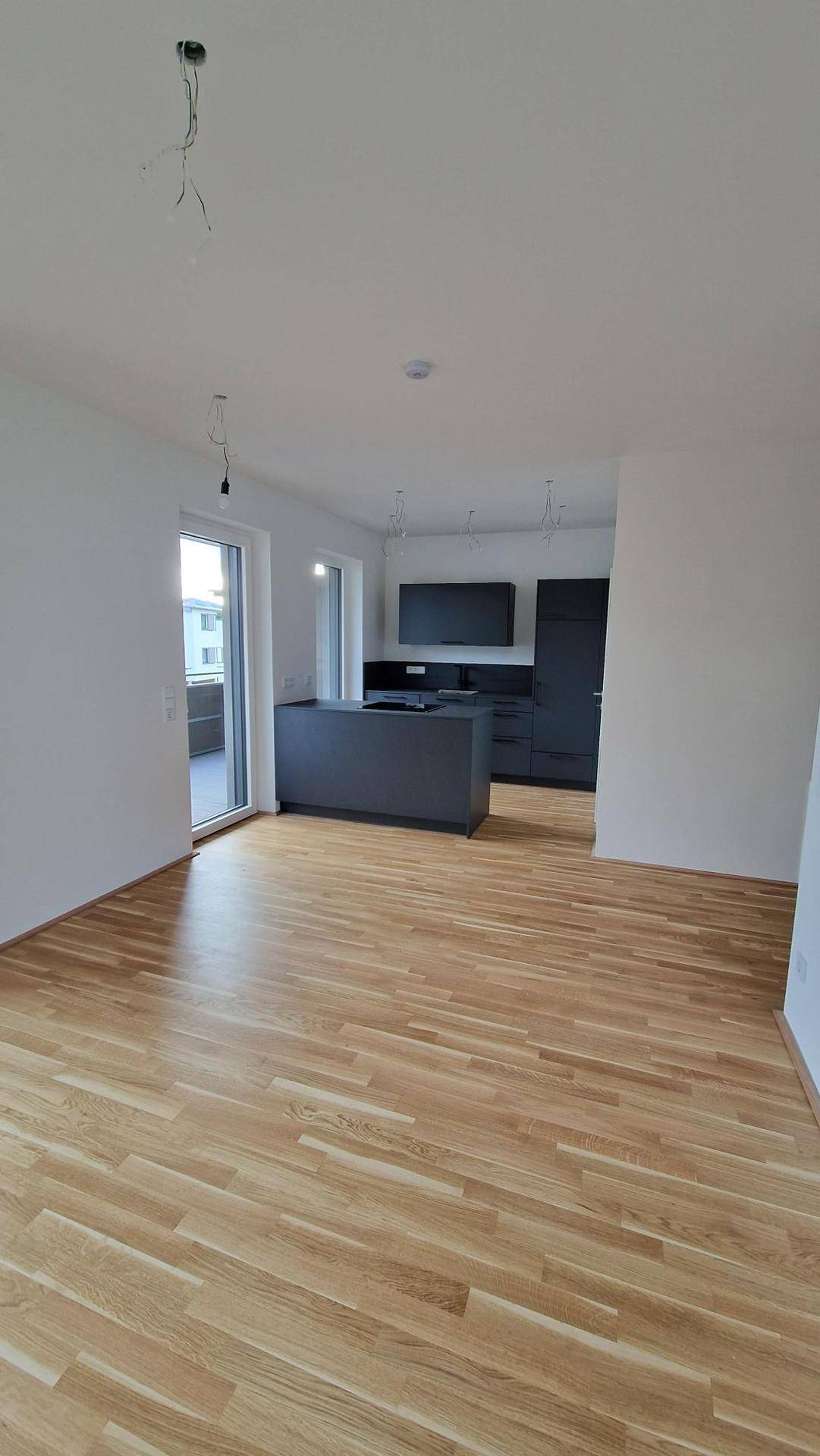 Offener Wohnbereich mit Holzboden und Blick auf die moderne graue Küche, mit Balkonzugang.