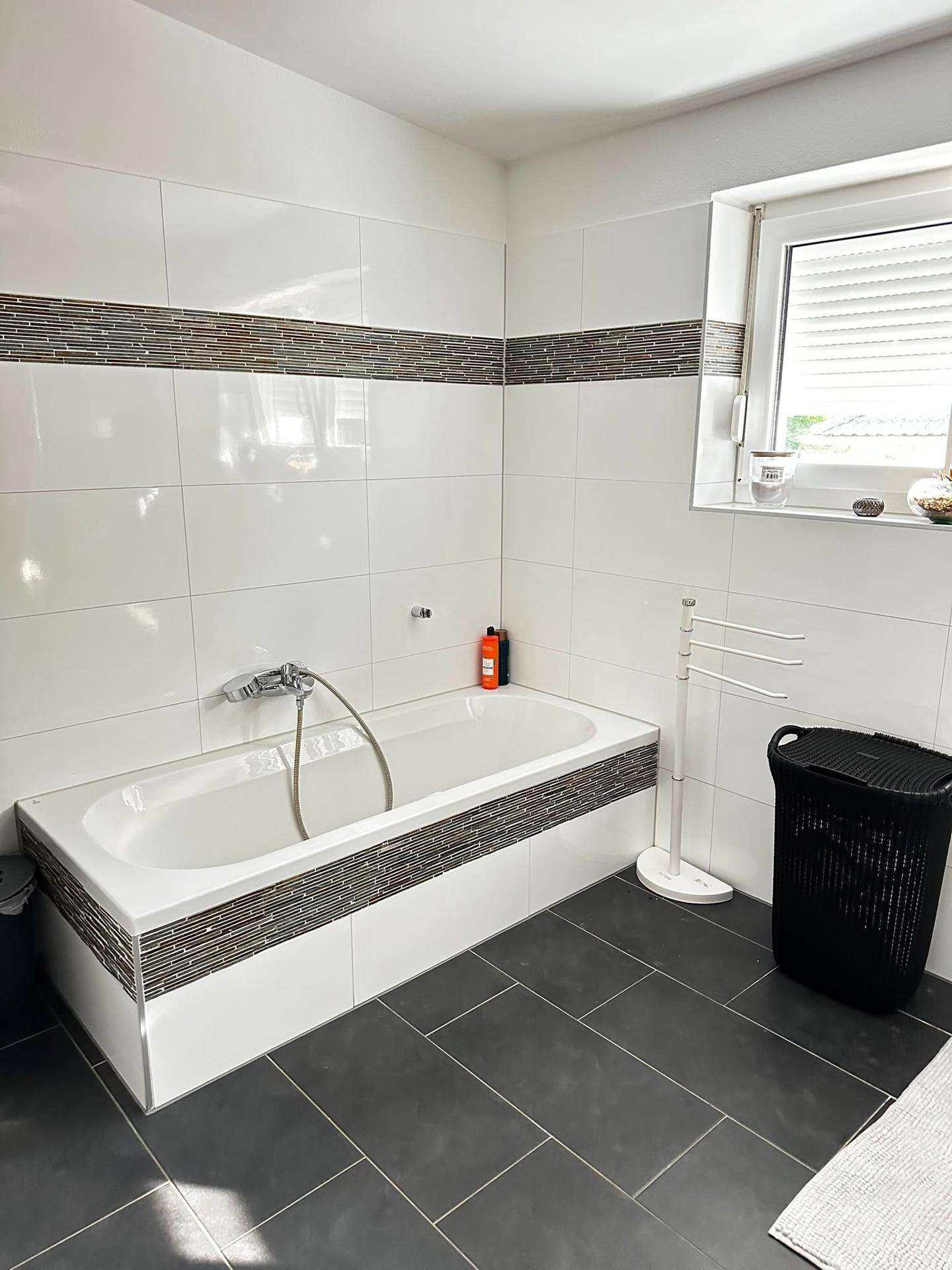 Helles Badezimmer mit Badewanne, dunklen Bodenfliesen und Fenster.