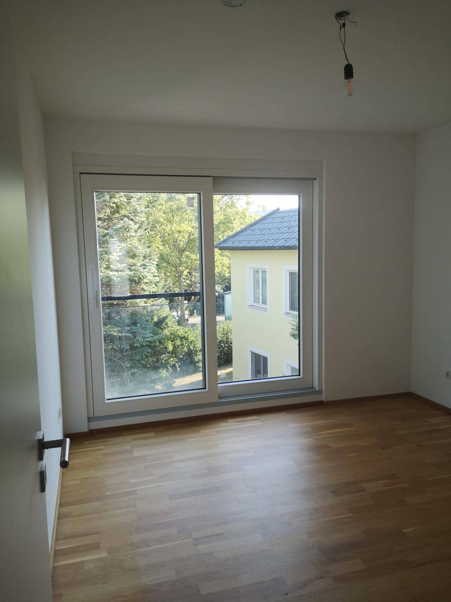 Schlafzimmer mit Holzboden und großem Fenster, das einen Blick auf ein Nachbargebäude bietet.