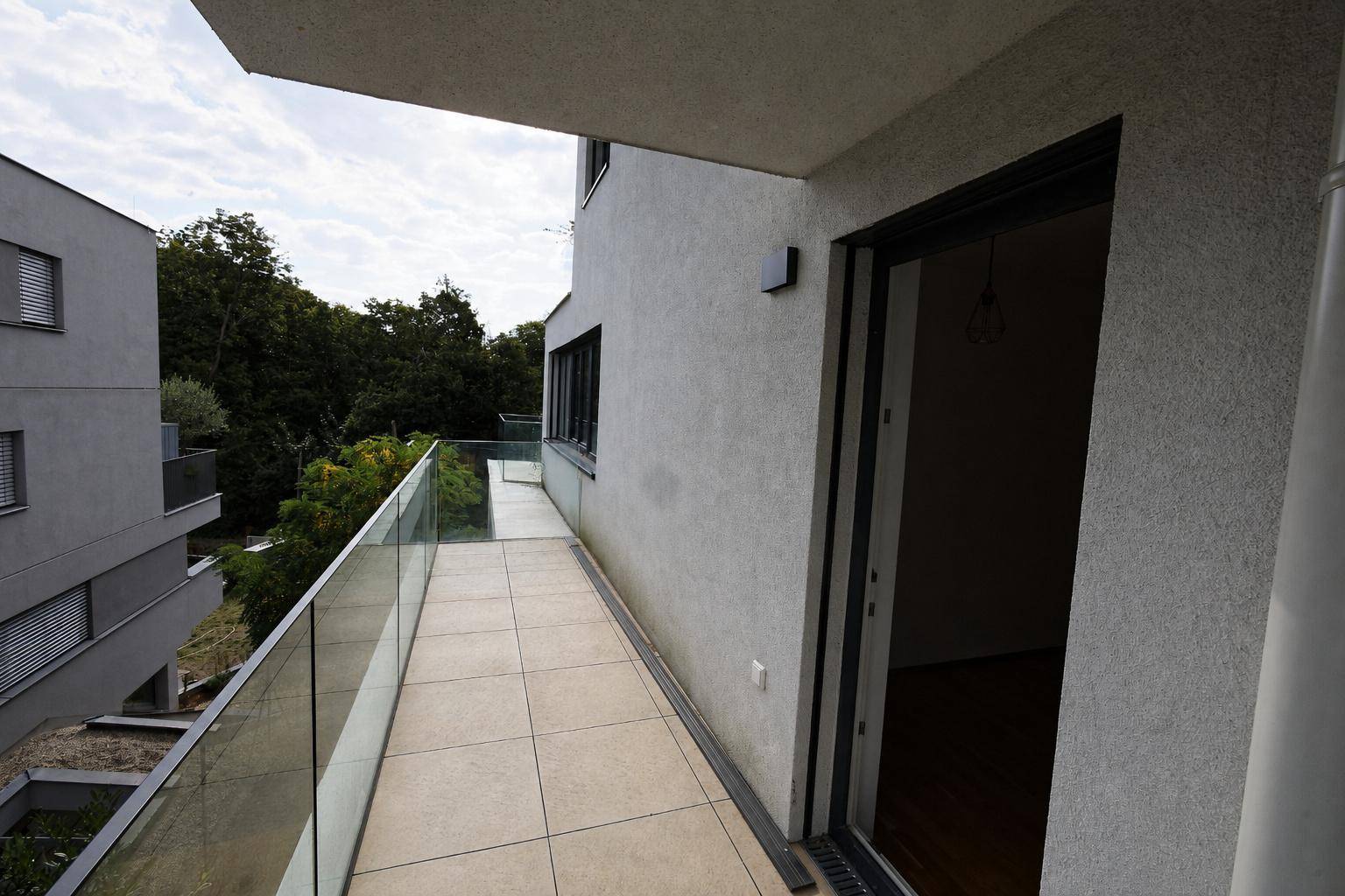 Langer, schmaler Balkon mit Glasgeländer und Blick auf die umliegenden Gebäude und Bäume.