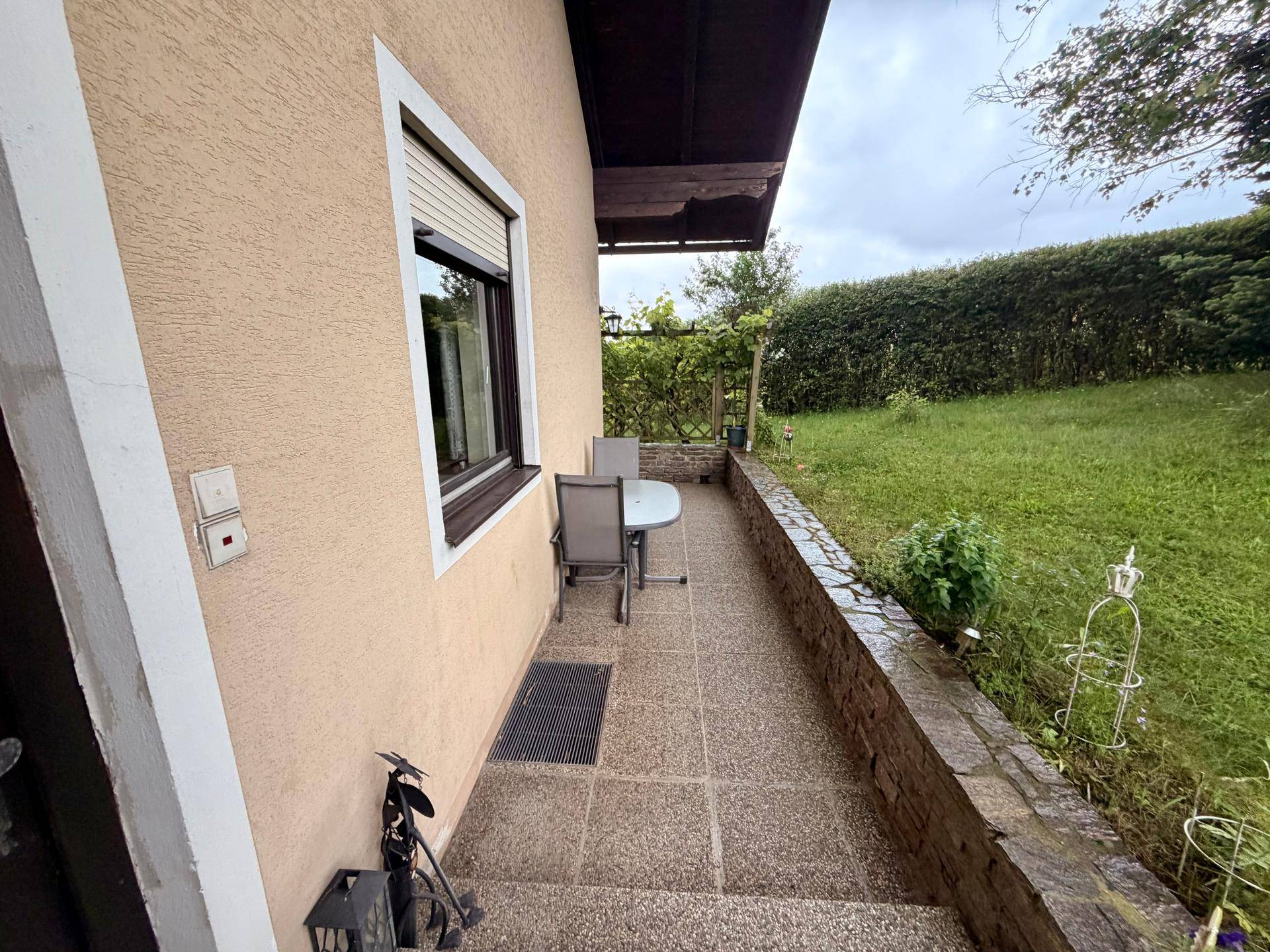Kleiner gepflasterter Terrassenbereich mit Sitzgelegenheit, umgeben von Grün und einer Hecke.