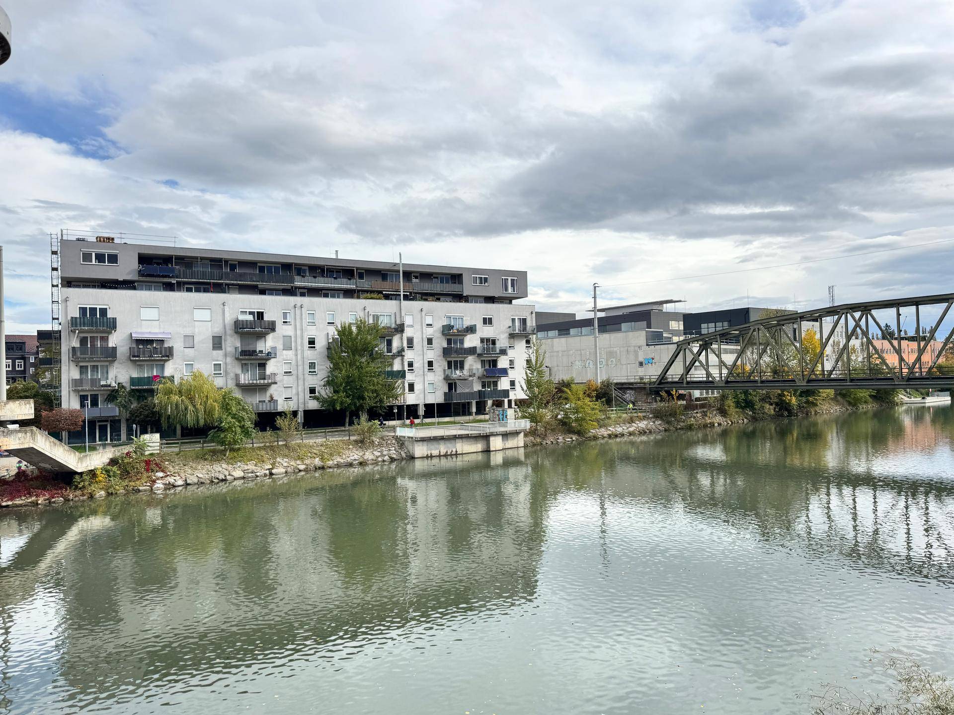 Wohngebäude am Flussufer mit Blick auf die umliegende Landschaft und eine Eisenbahnbrücke unter bewölktem Himmel.
