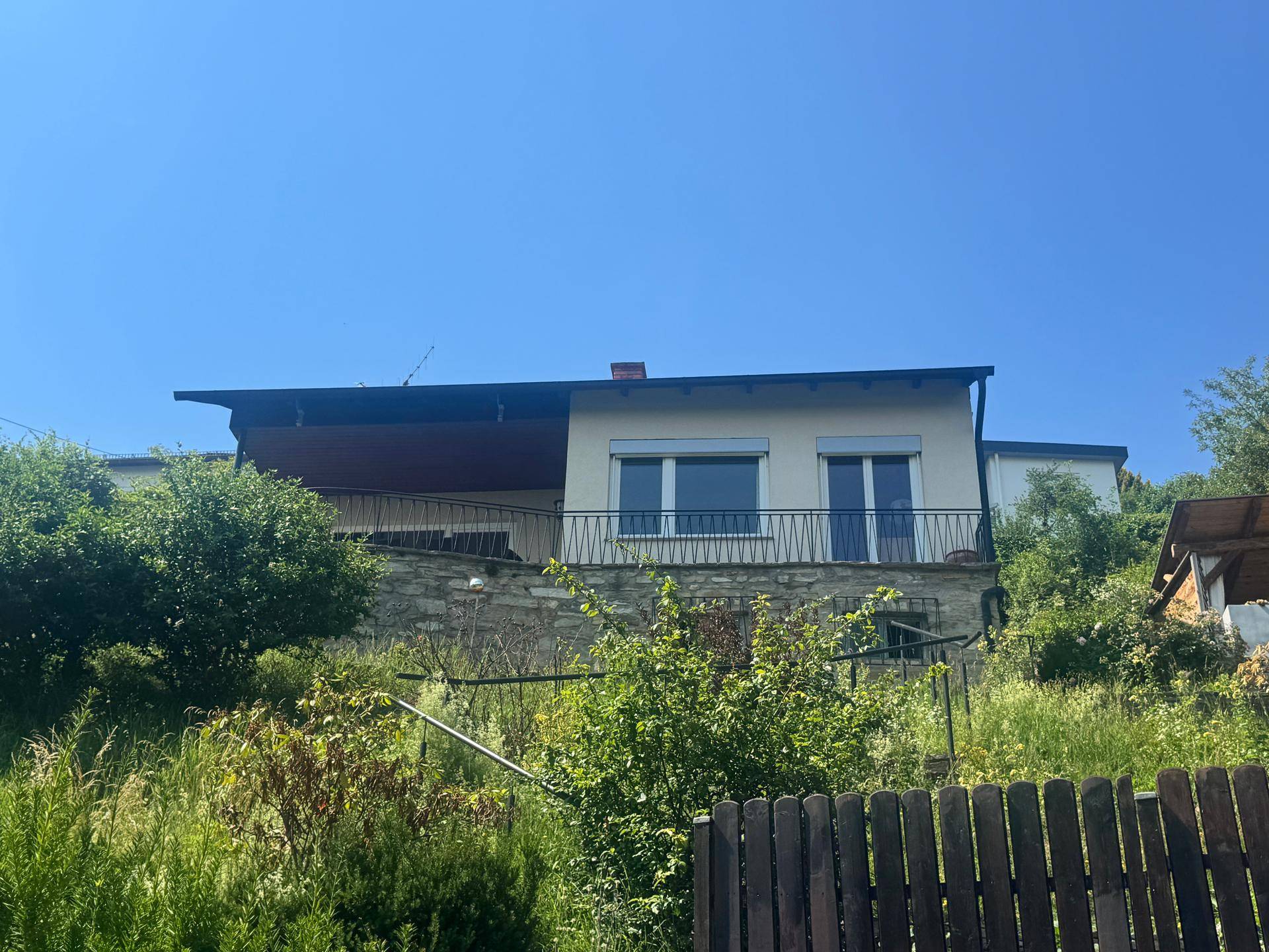 Ansicht des Hauses mit Balkon und Rollläden, umgeben von üppiger Vegetation an einem sonnigen Tag.