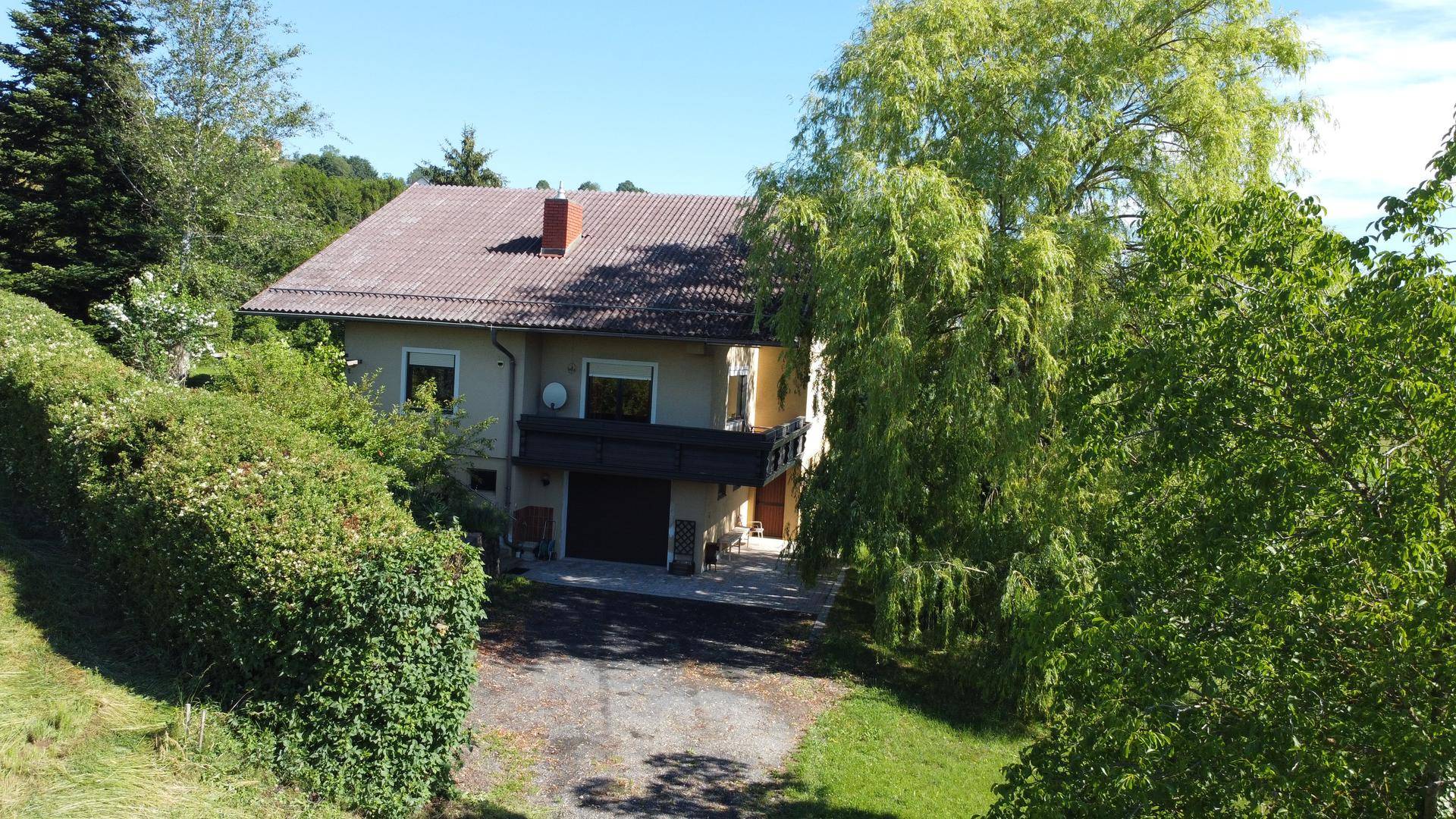 Frontansicht des Hauses mit Balkon und Zufahrt, umgeben von üppiger Vegetation.