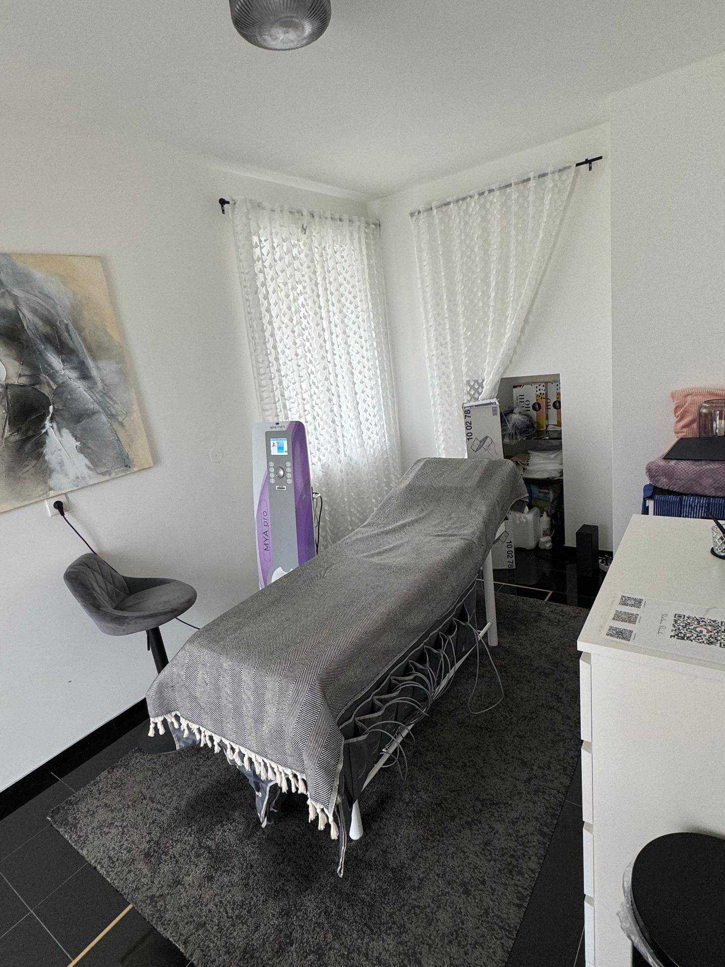 Behandlungsraum mit Liege, medizinischem Gerät und dunklem Teppichboden, hell und funktional.