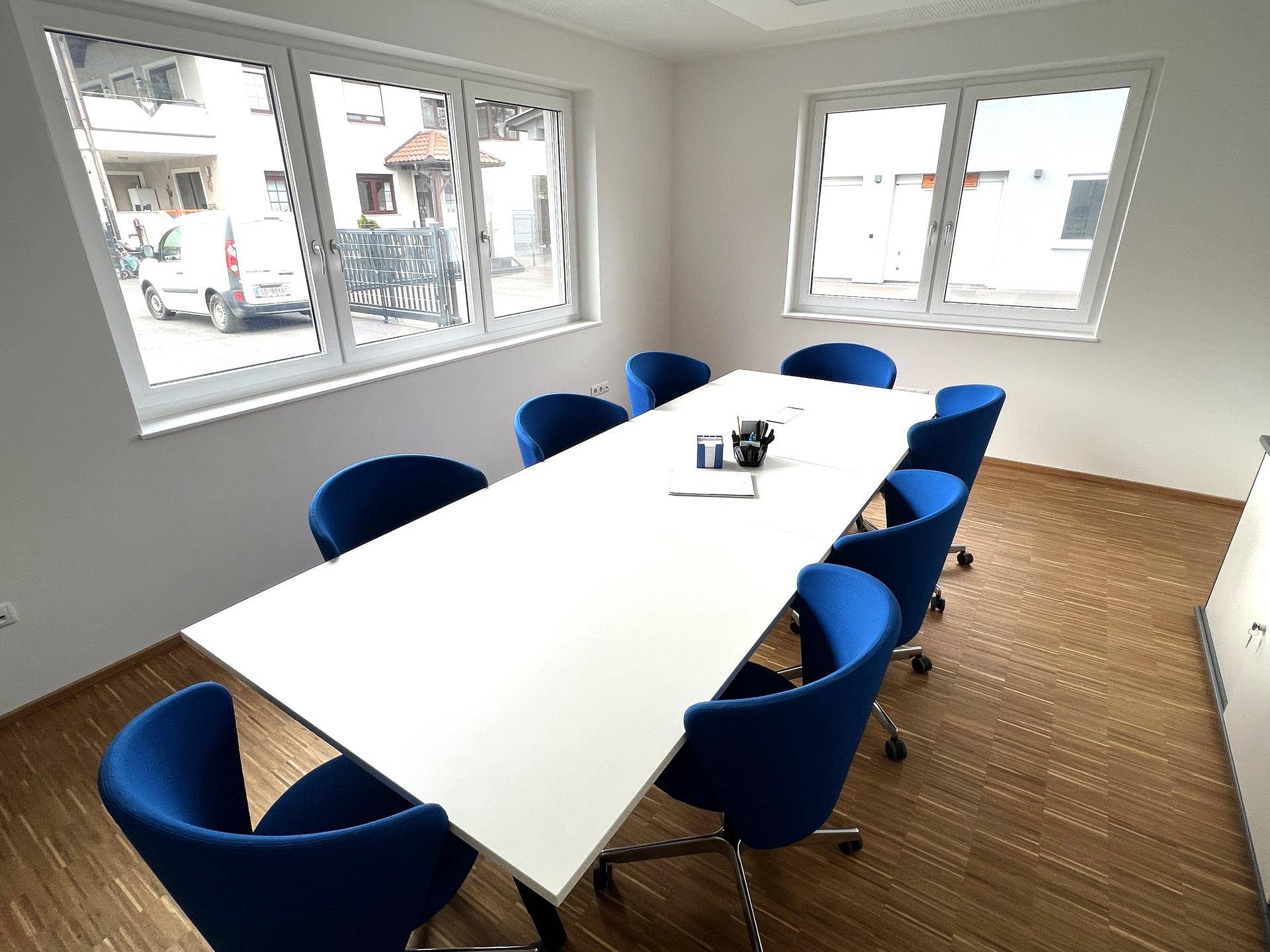 Moderner Konferenzraum mit großem weißen Tisch und blauen Stühlen, viel Tageslicht durch große Fenster.