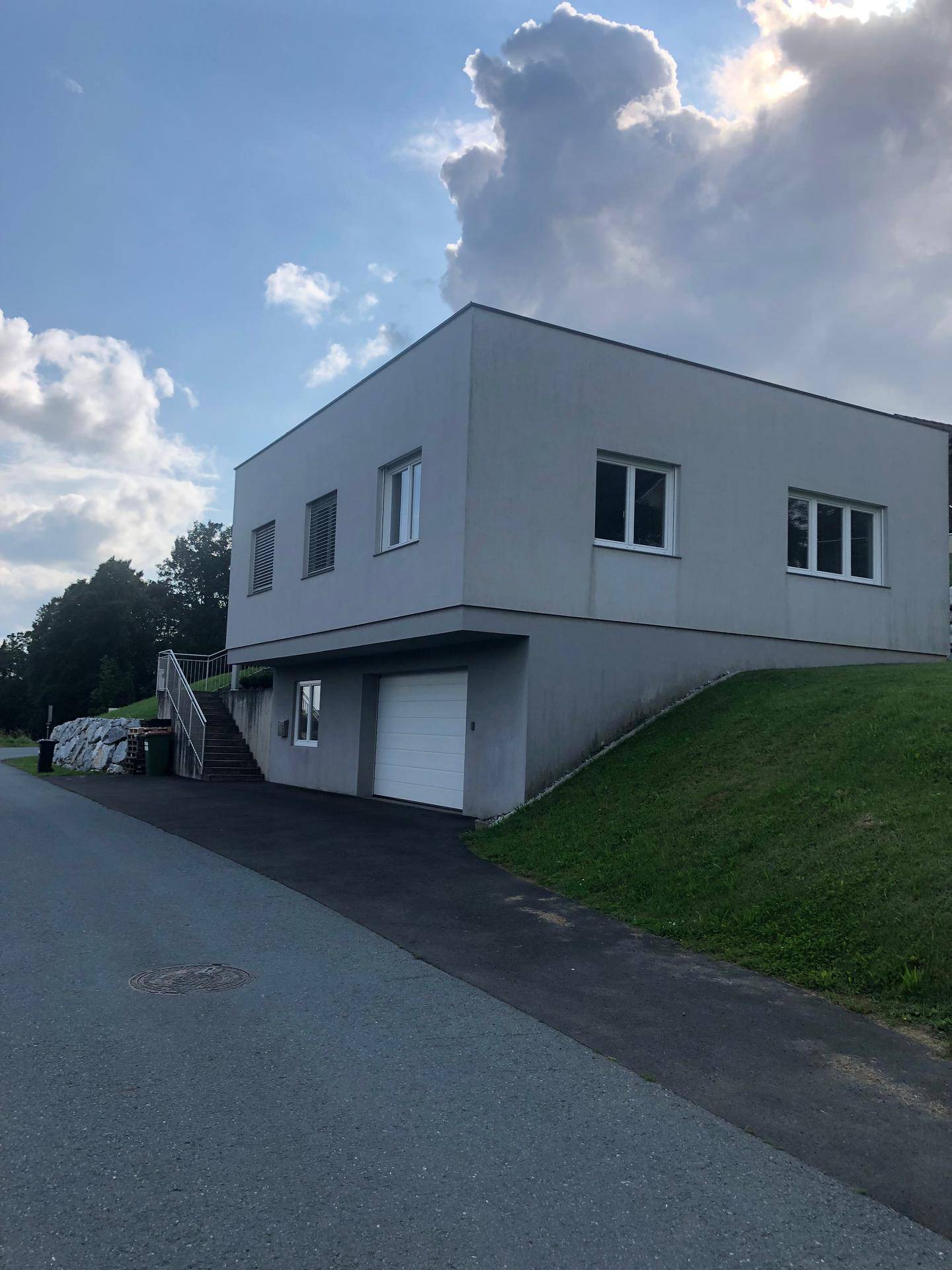 Modernes Gebäude mit schlichter Fassade, Zufahrt und Hanglage unter bewölktem Himmel.