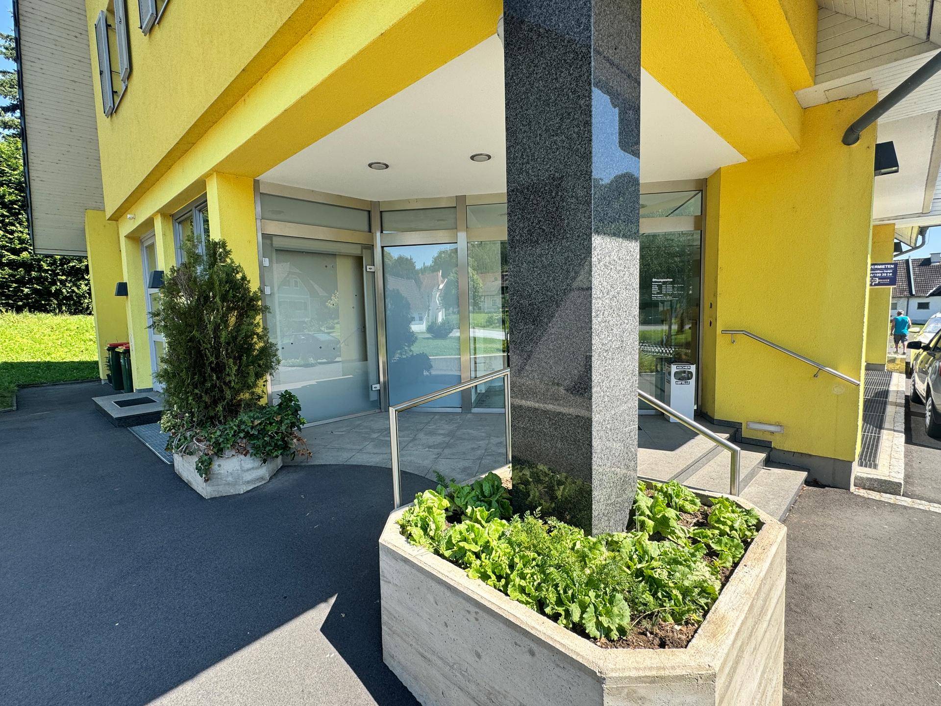 Eingangsbereich eines gelben Gebäudes mit Granitsäule, automatischen Türen und Pflanzenkübeln.
