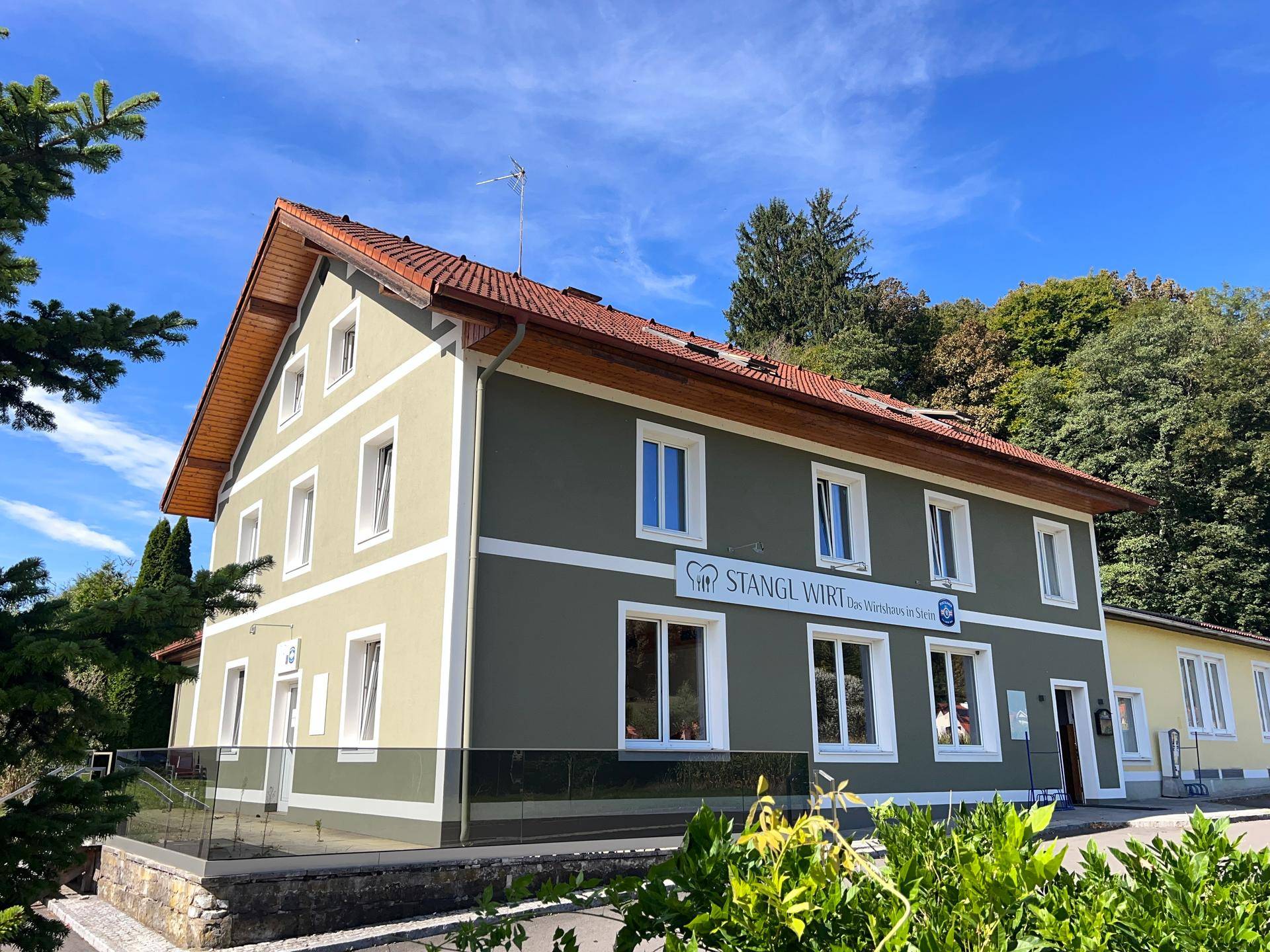 Traditionelles Gebäude mit grüner Fassade und rotem Ziegeldach, umgeben von Bäumen und einem Parkplatz.