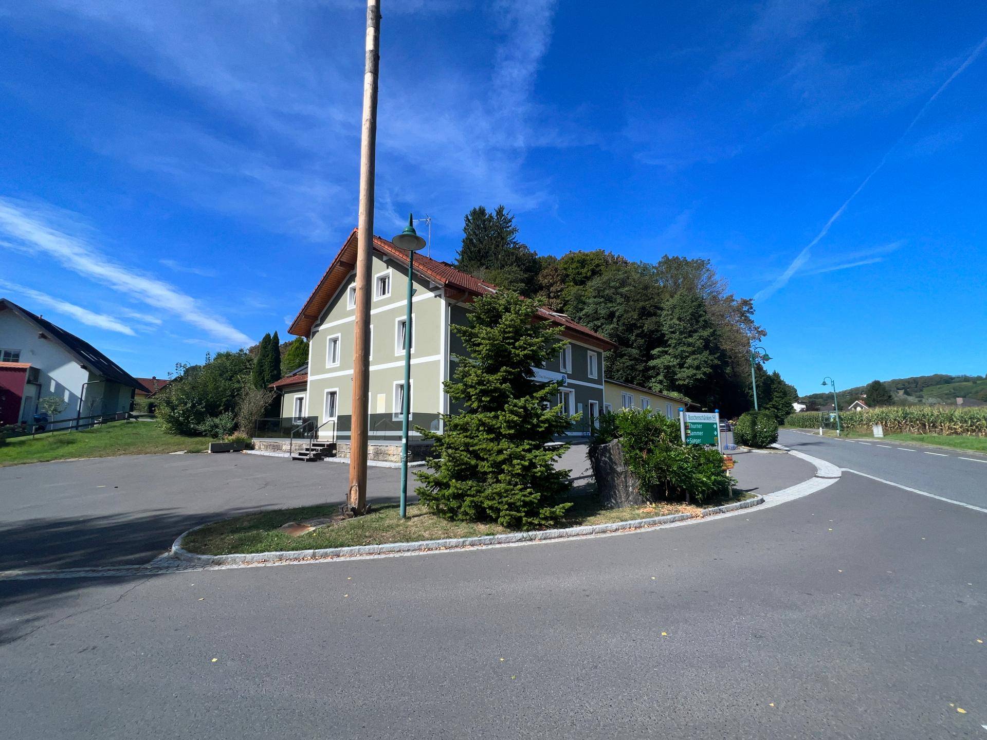 Panoramablick auf das Gebäude mit Maibaum, umgeben von ländlicher Umgebung und einer Straße.