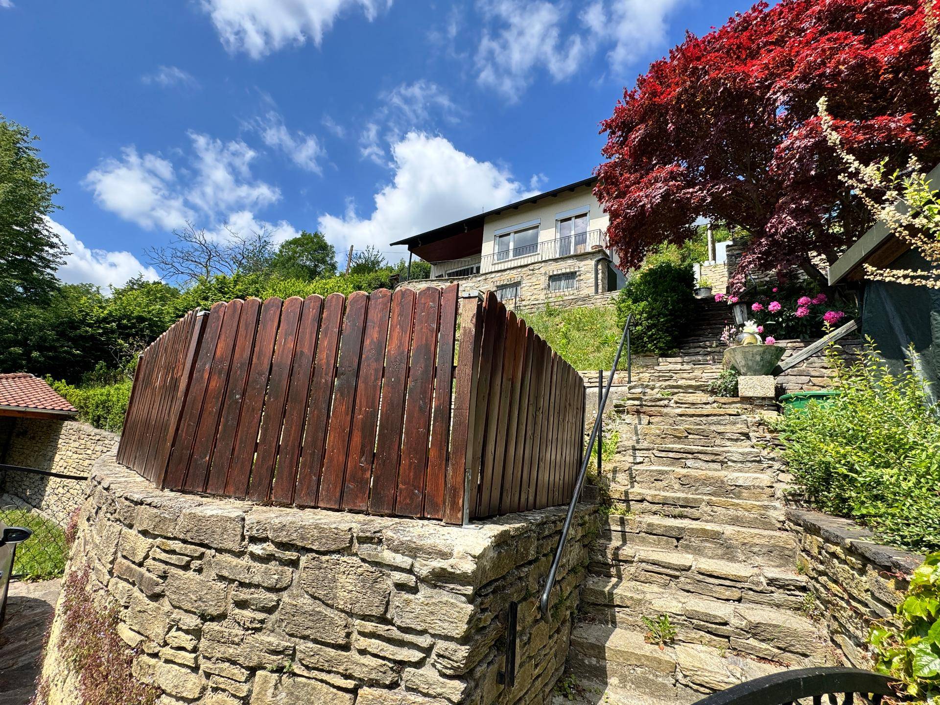 Das Haus auf dem Hügel, umgeben von einer Steinmauer und Holzumzäunung, mit roten Bäumen im Vordergrund.
