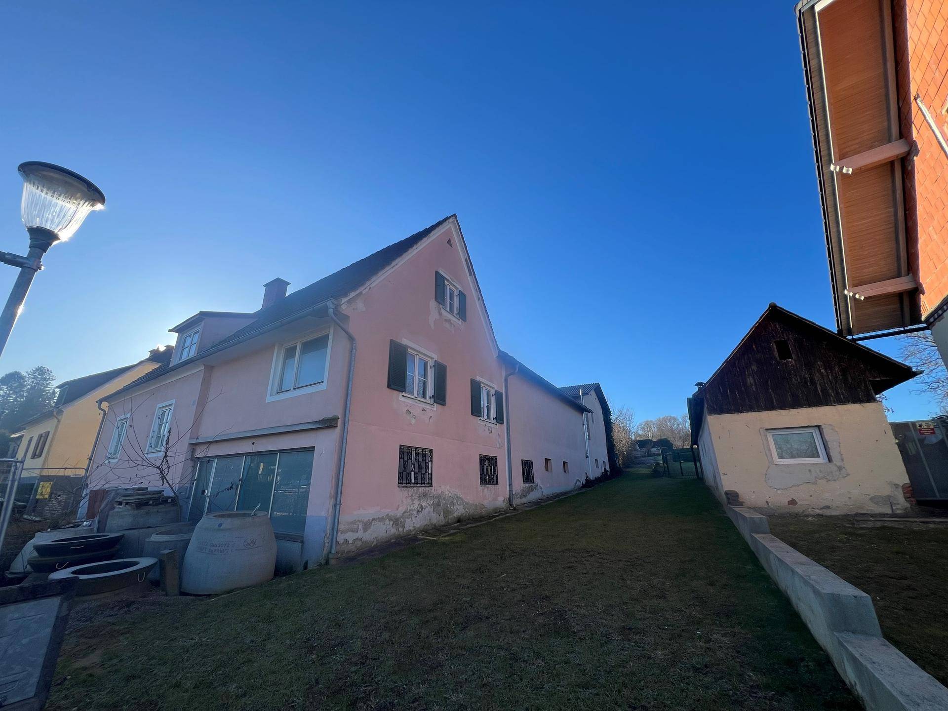 Seitenansicht des rosa Hauses mit angrenzendem Nebengebäude und einem Hang im Hintergrund.