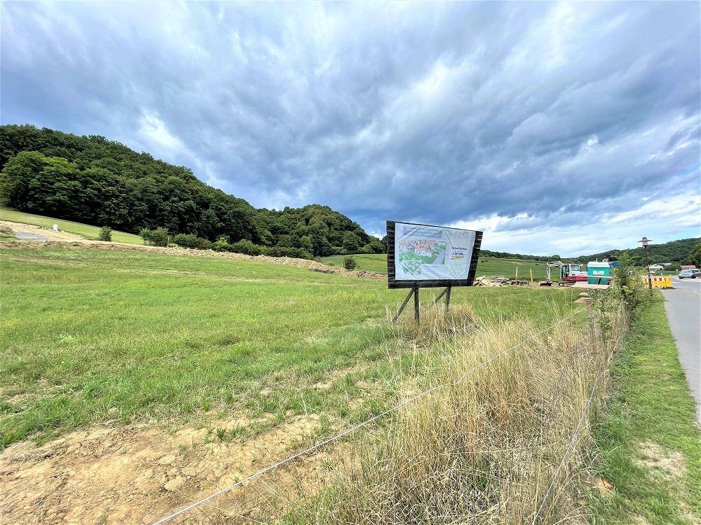 Grünfläche mit Informationsschild und Blick auf bewaldete Hügel unter bewölktem Himmel.