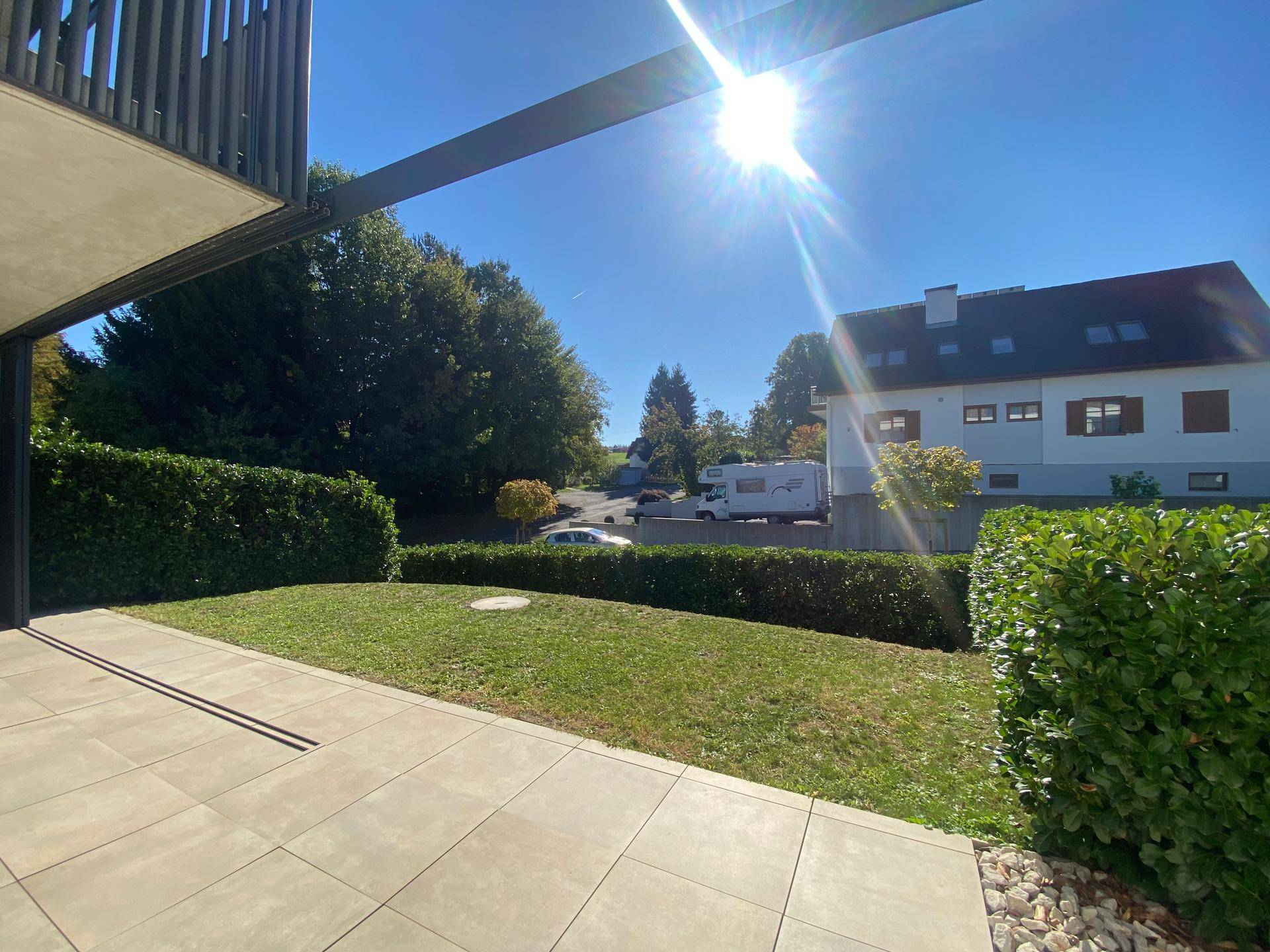 Sonniger Außenbereich mit Terrasse und Rasenfläche, umgeben von Hecken und Bäumen.