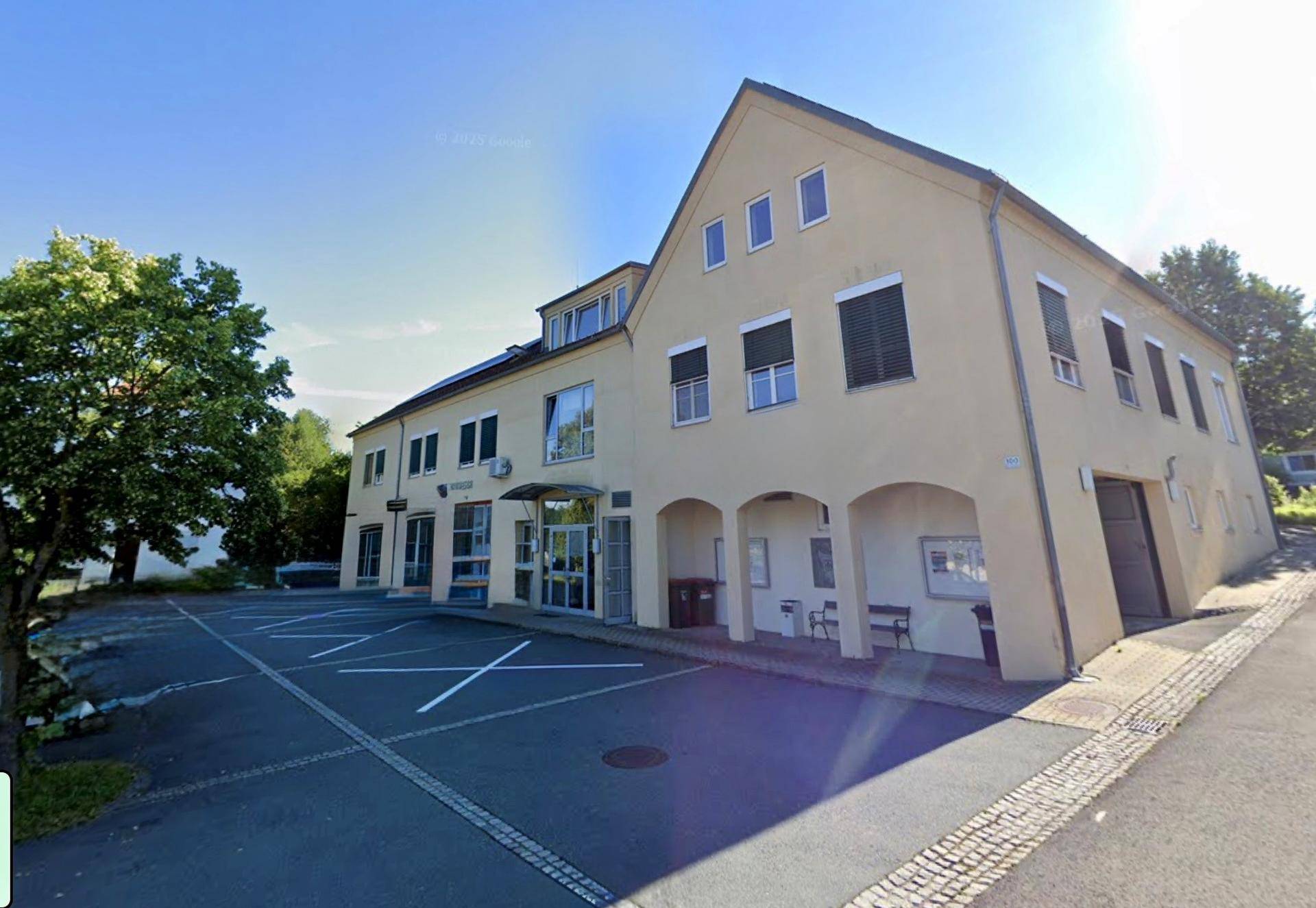 Gebäudeansicht mit Parkplätzen und Bäumen an einem sonnigen Tag.