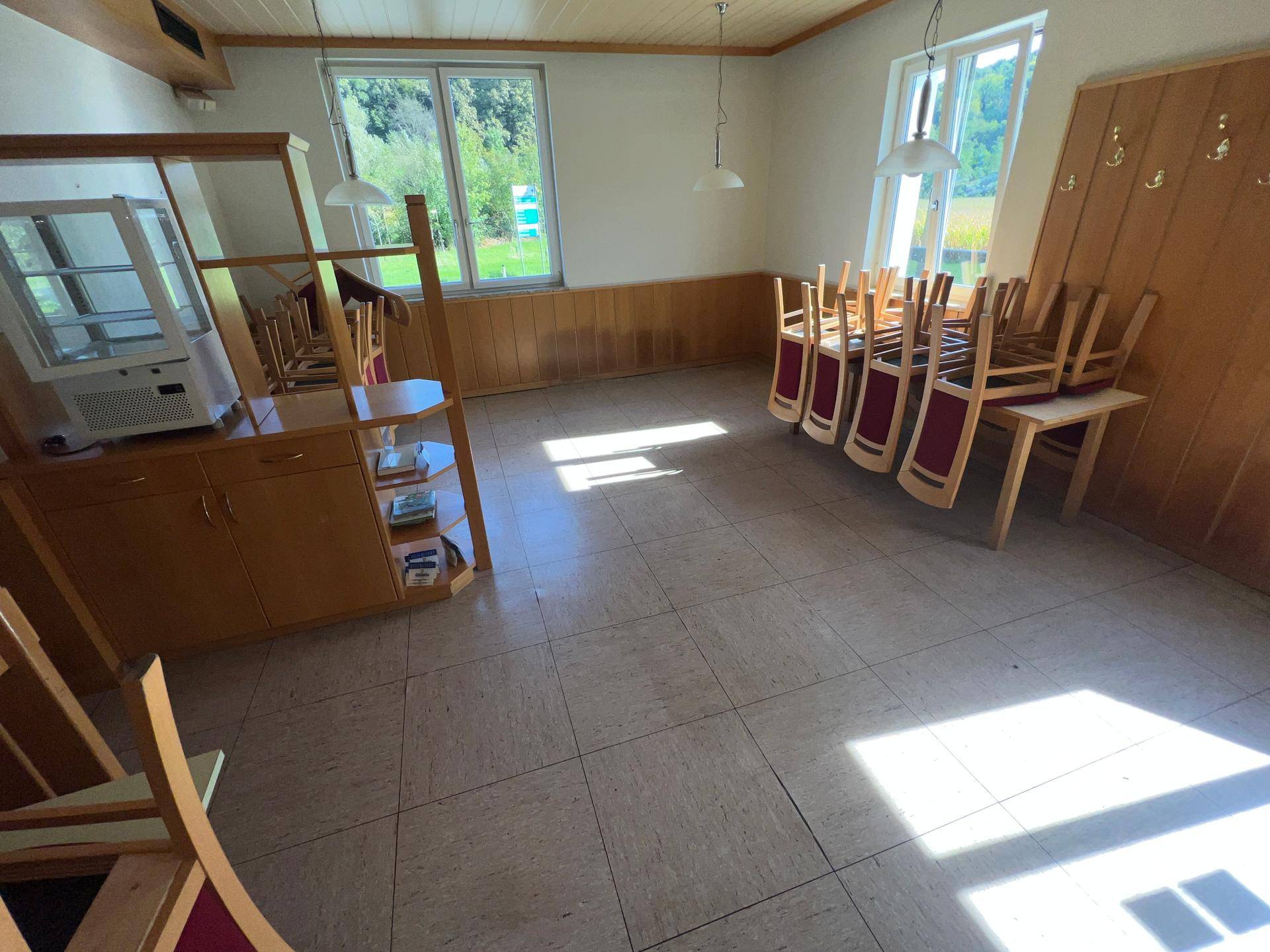 Speisesaal mit gestapelten Stühlen, Vitrine und großen Fenstern, bereit für Veranstaltungen.