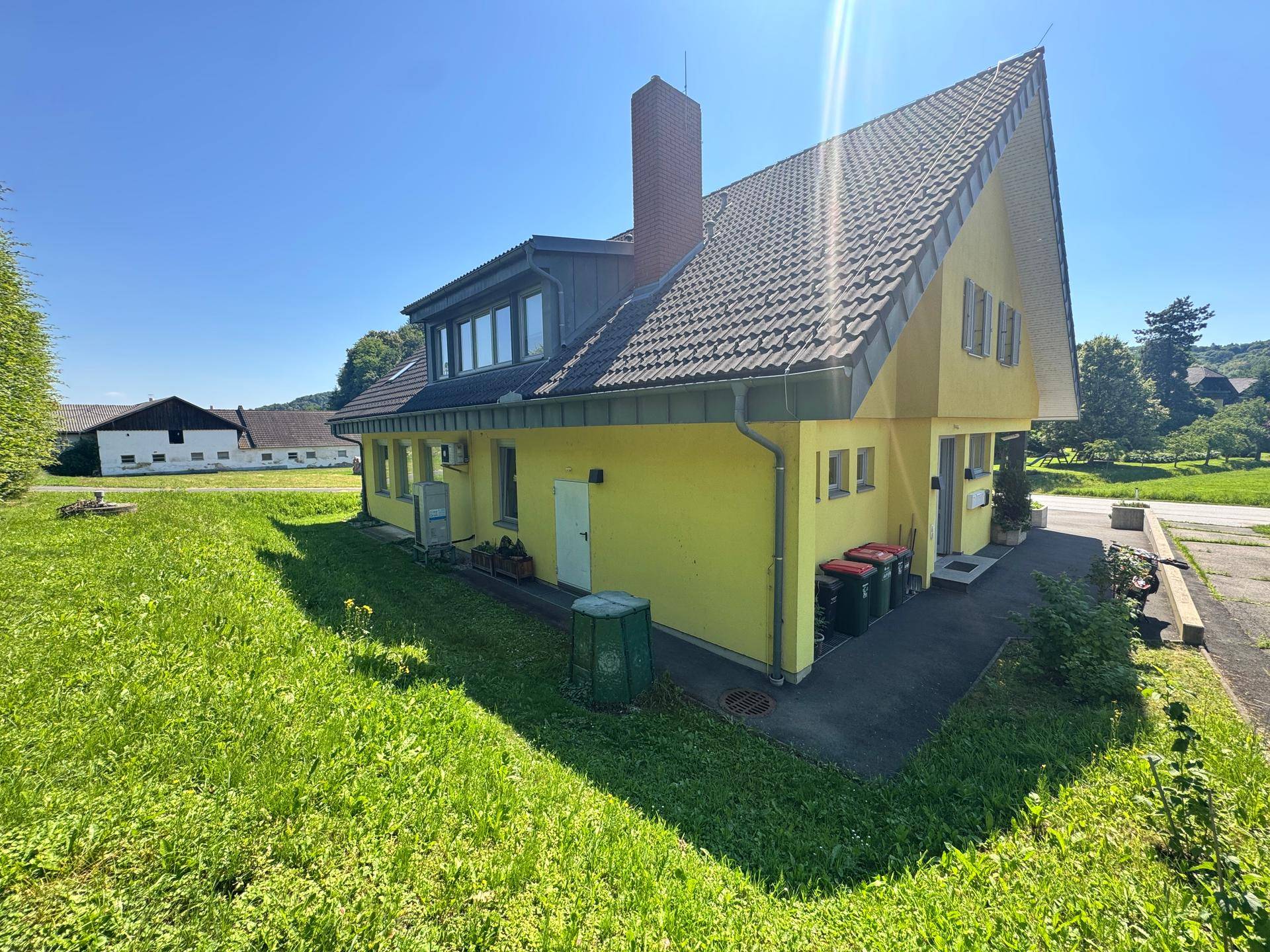 Rückansicht des gelben Hauses mit Schornstein, Satteldach und Mülltonnen im grünen Gartenbereich.