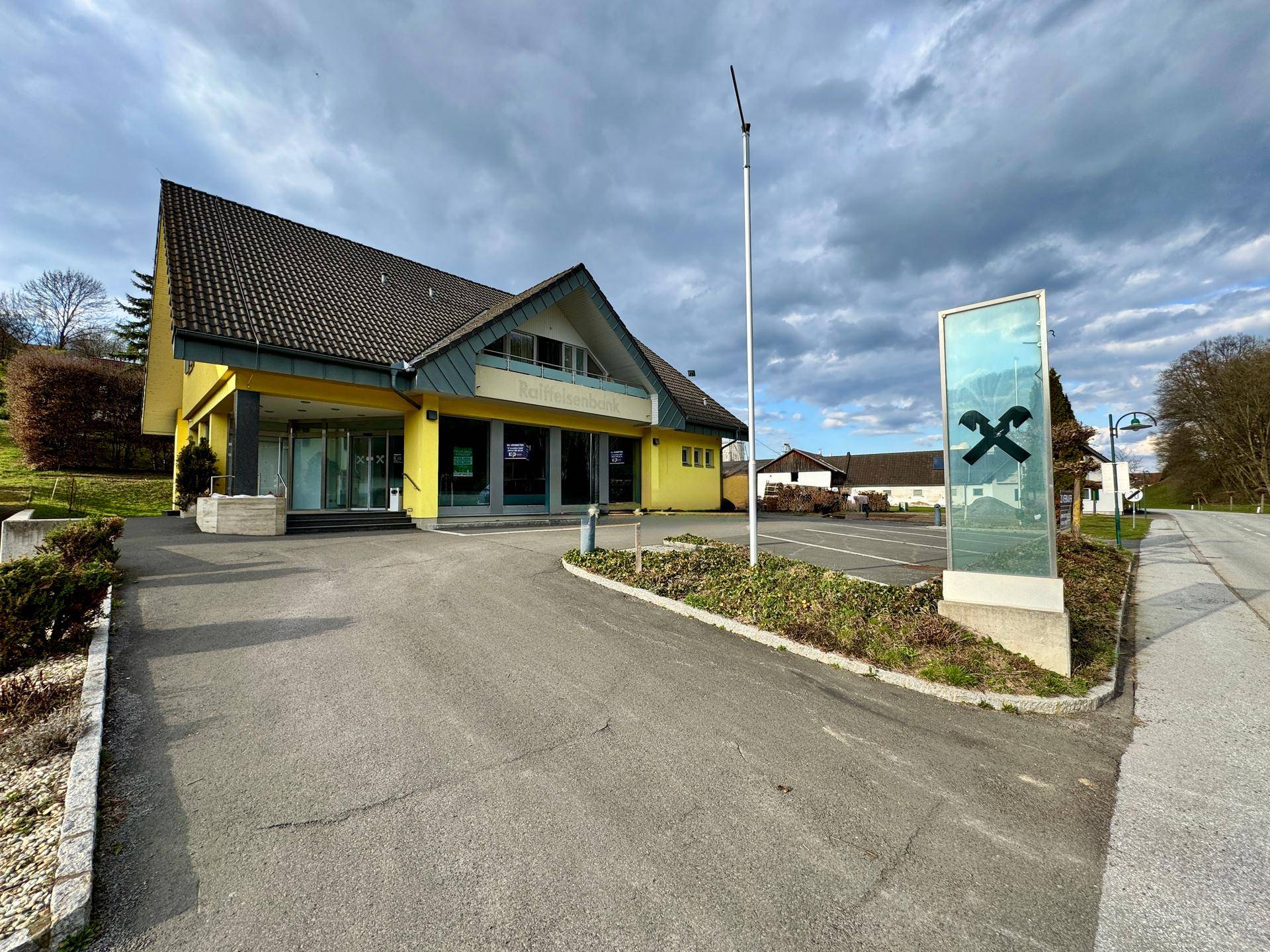 Gelbes Gebäude mit Satteldach und großem Parkplatz unter bewölktem Himmel.
