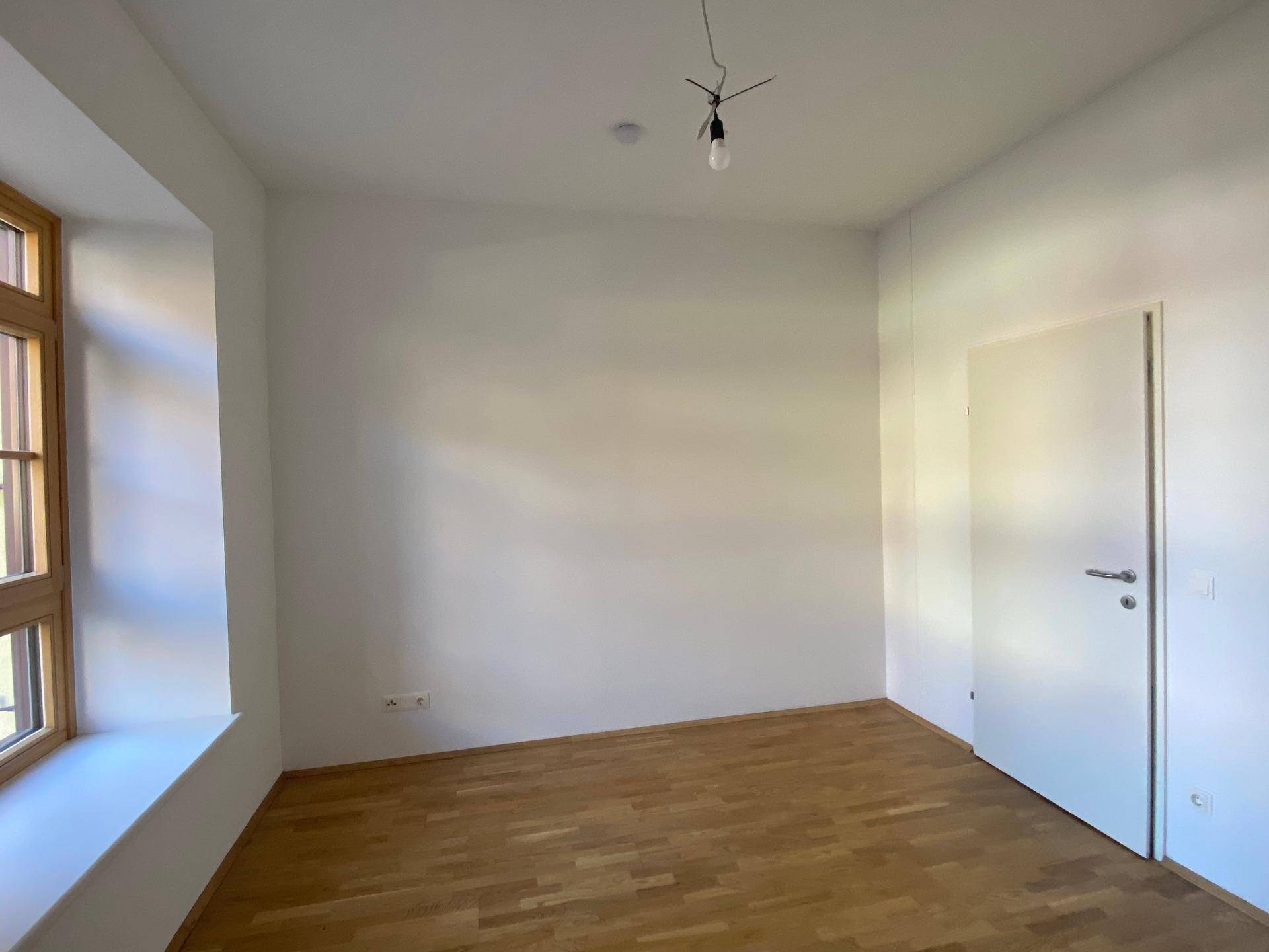 Leeres Zimmer mit hellem Parkettboden, weißen Wänden und einem Fenster, bereit zur Einrichtung.