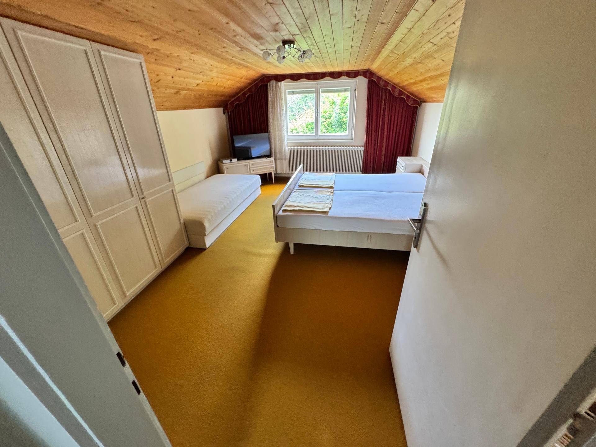 Schlafzimmer mit Holzdecke, Teppichboden und zwei Betten unter einem Fenster.