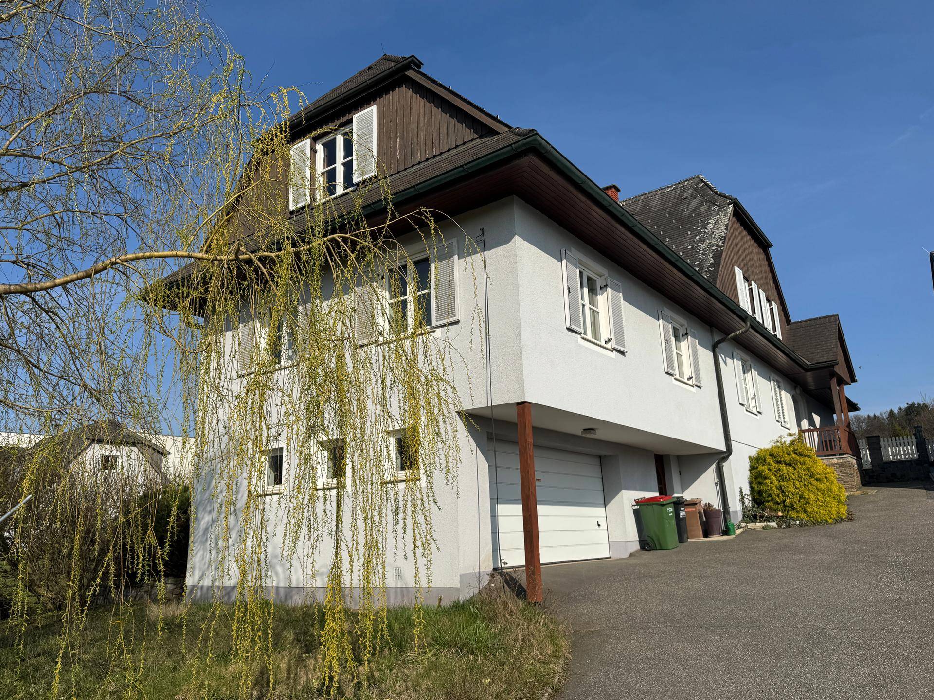 Seitenansicht des Hauses mit weißer Fassade, Garage und einem blühenden Baum im Vordergrund.