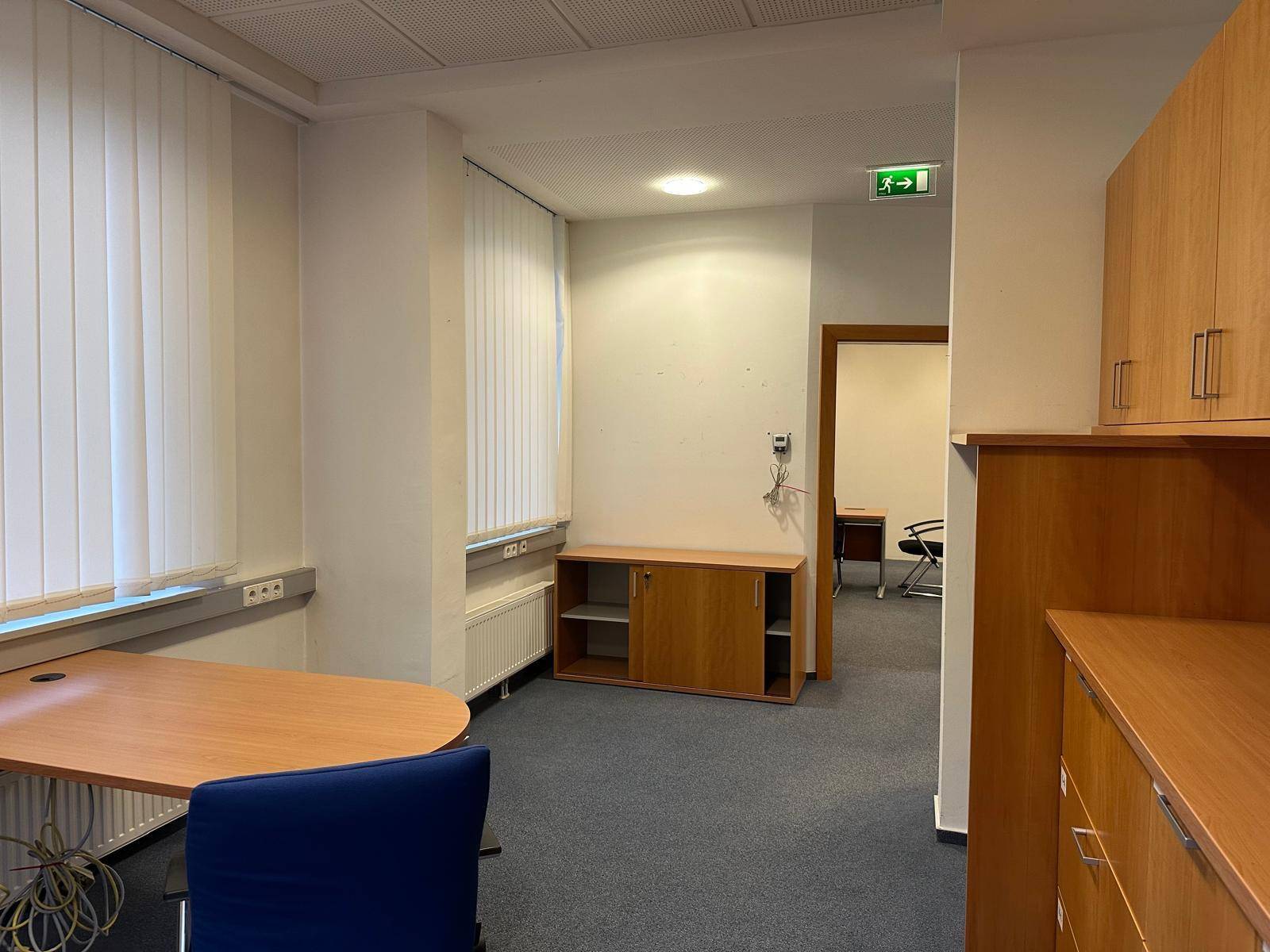 Büro mit Teppichboden, Fenster und Zugang zu einem weiteren Raum.