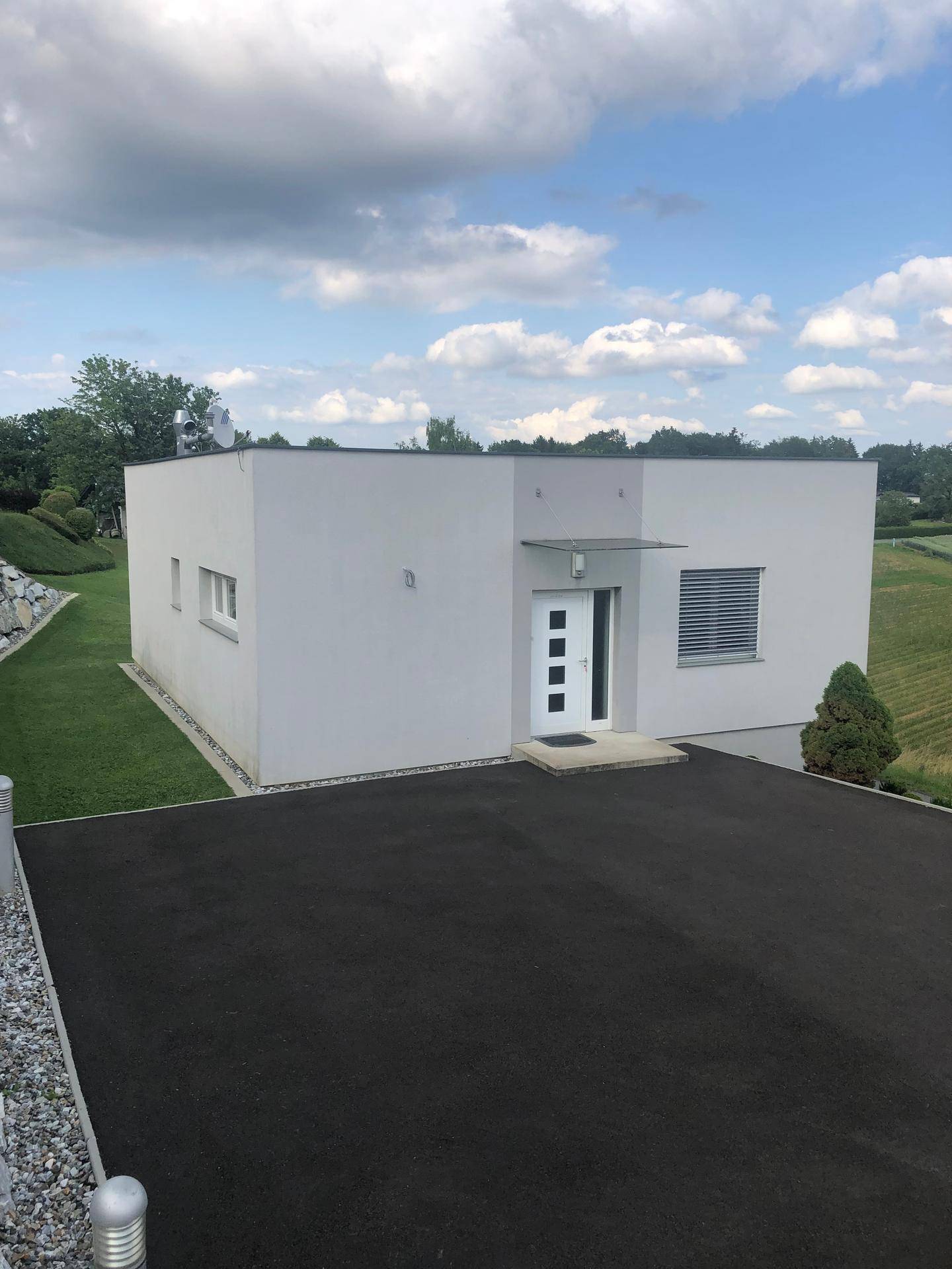 Modernes Gebäude mit flachem Dach, umgeben von Grünflächen und einer asphaltierten Zufahrt.