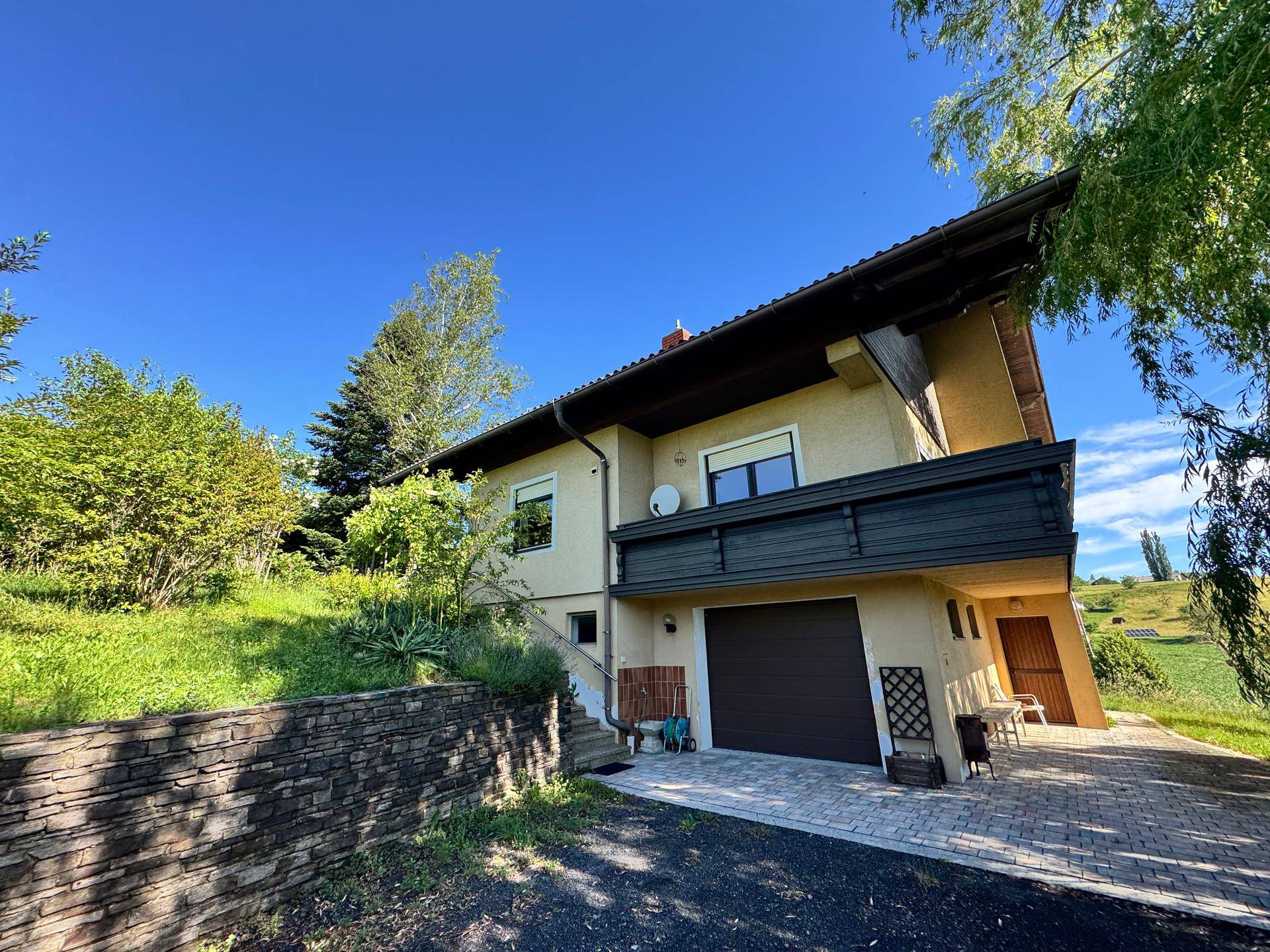 Haus mit Garage und Balkon, umgeben von einer Steinmauer und grüner Landschaft.