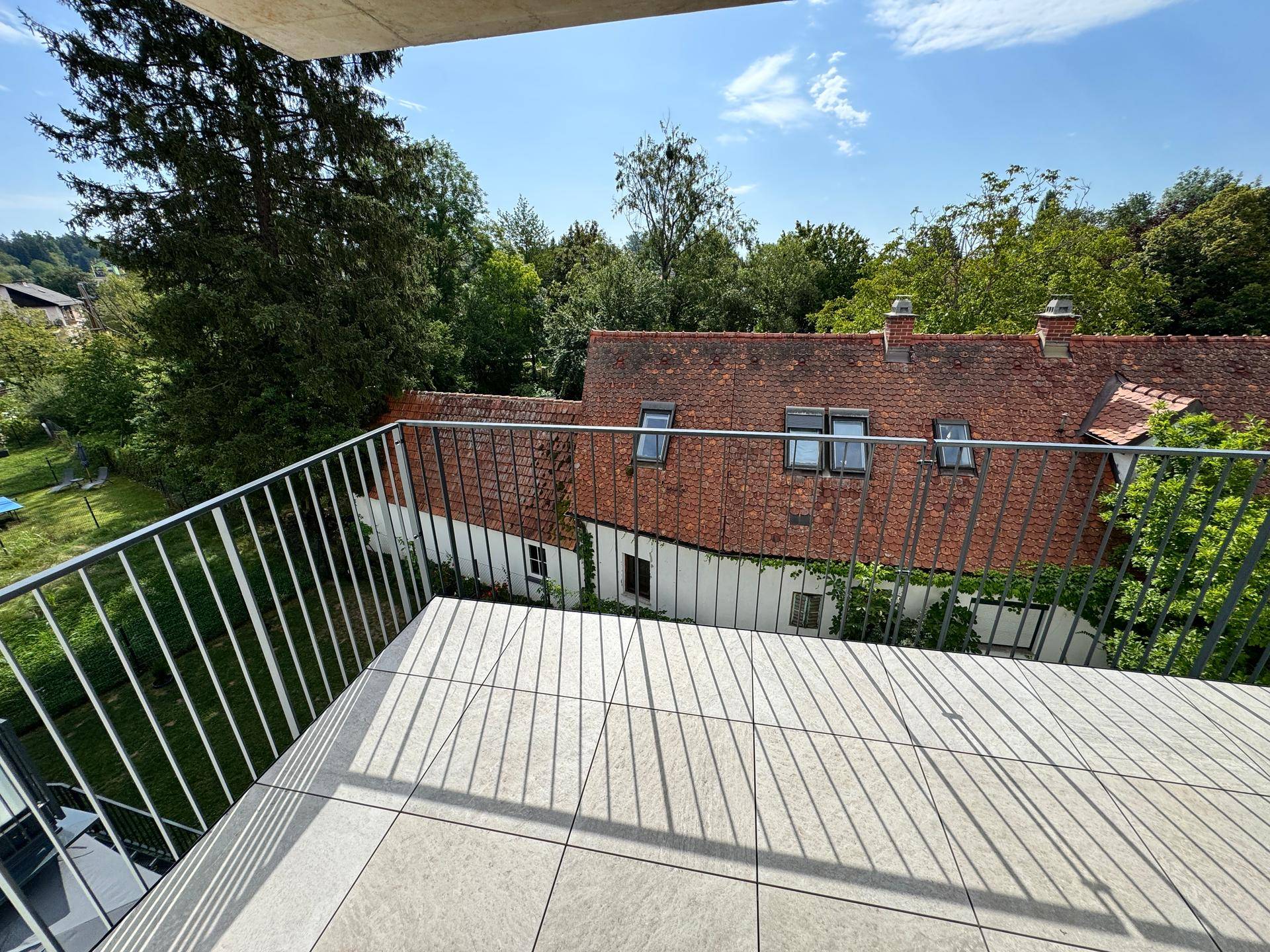 Balkon mit Fliesenboden und Blick auf die umliegende Natur und Dächer, sehr sonnig.