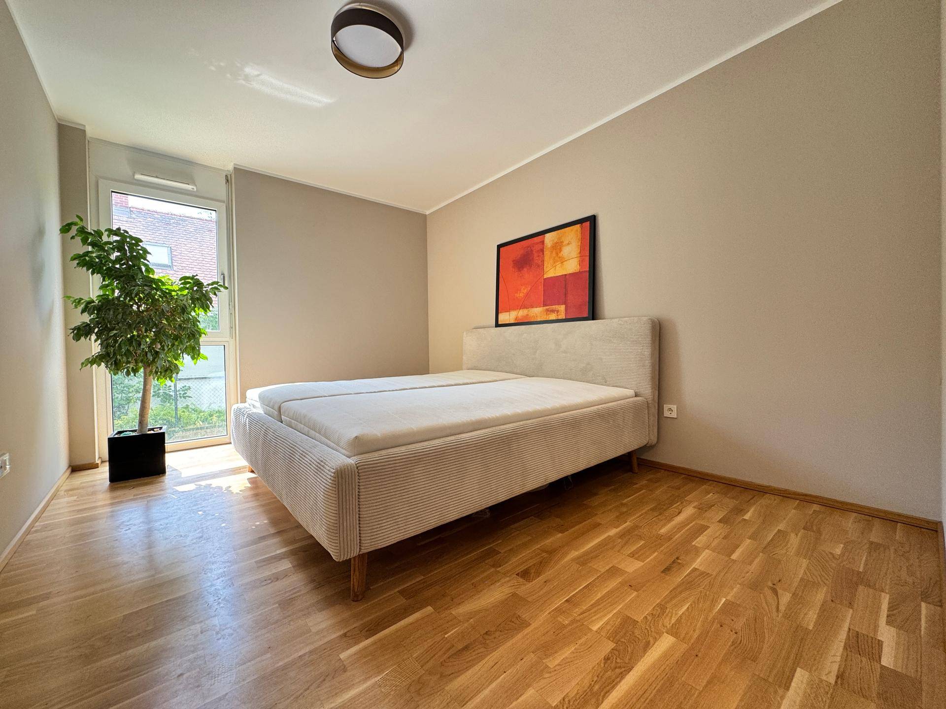Helles Schlafzimmer mit Doppelbett, Holzparkettboden und großem Fenster mit viel Tageslicht.
