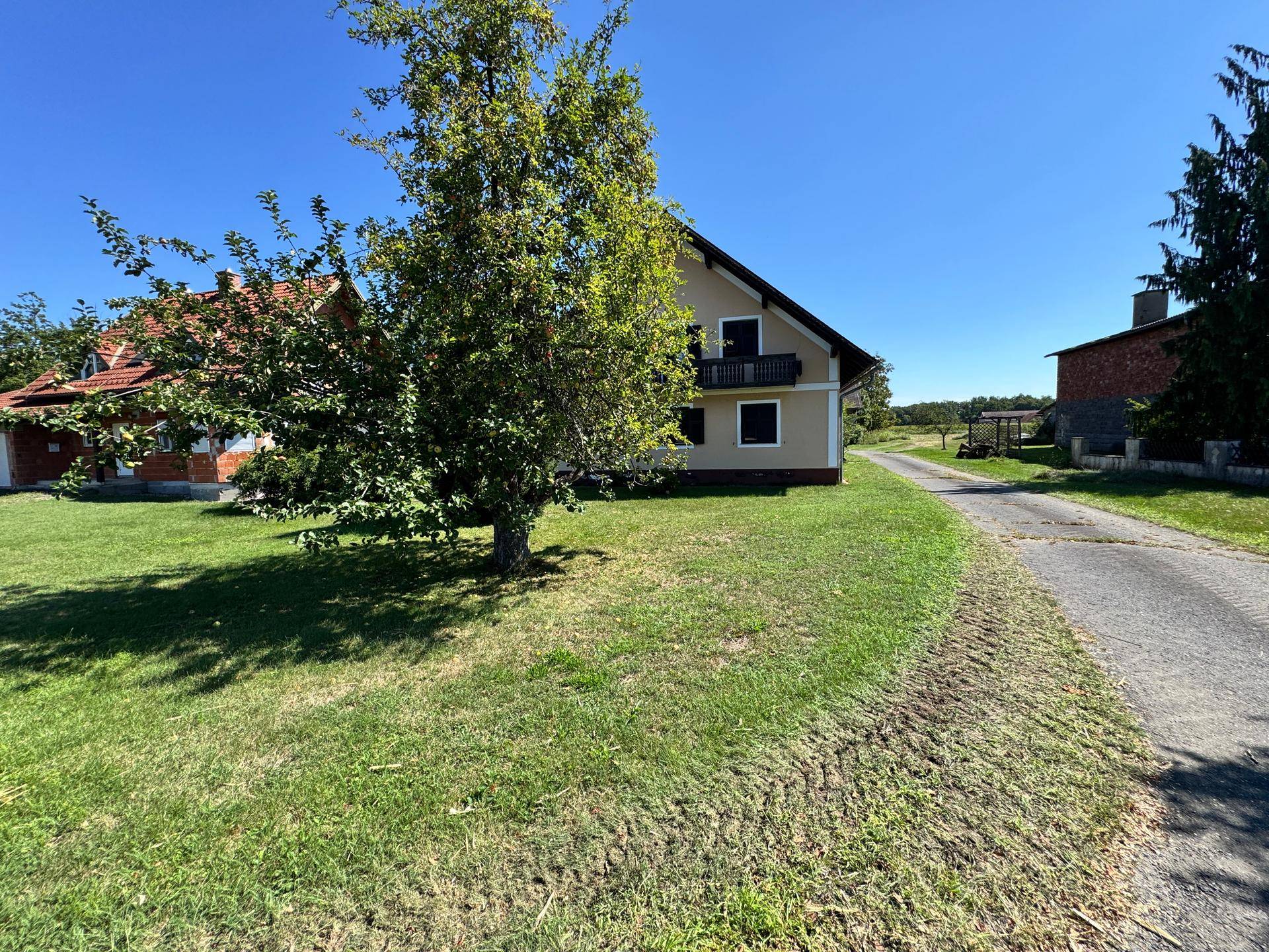 Haus mit Balkon, grüner Wiese und Zufahrtsweg in ländlicher Umgebung unter blauem Himmel.