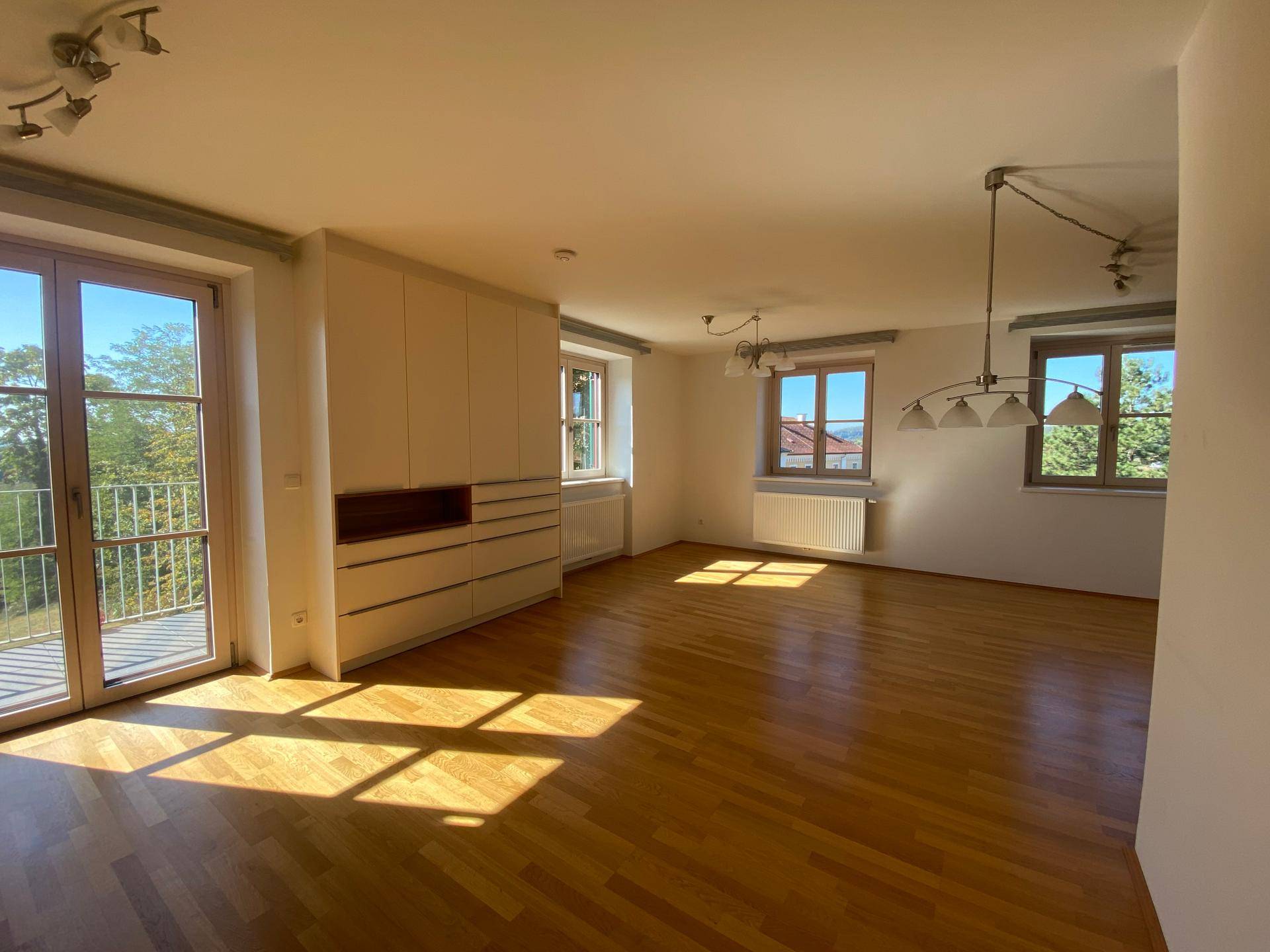 Geräumiges Wohnzimmer mit sonnigem Holzboden, großen Fenstern und Zugang zum Balkon.