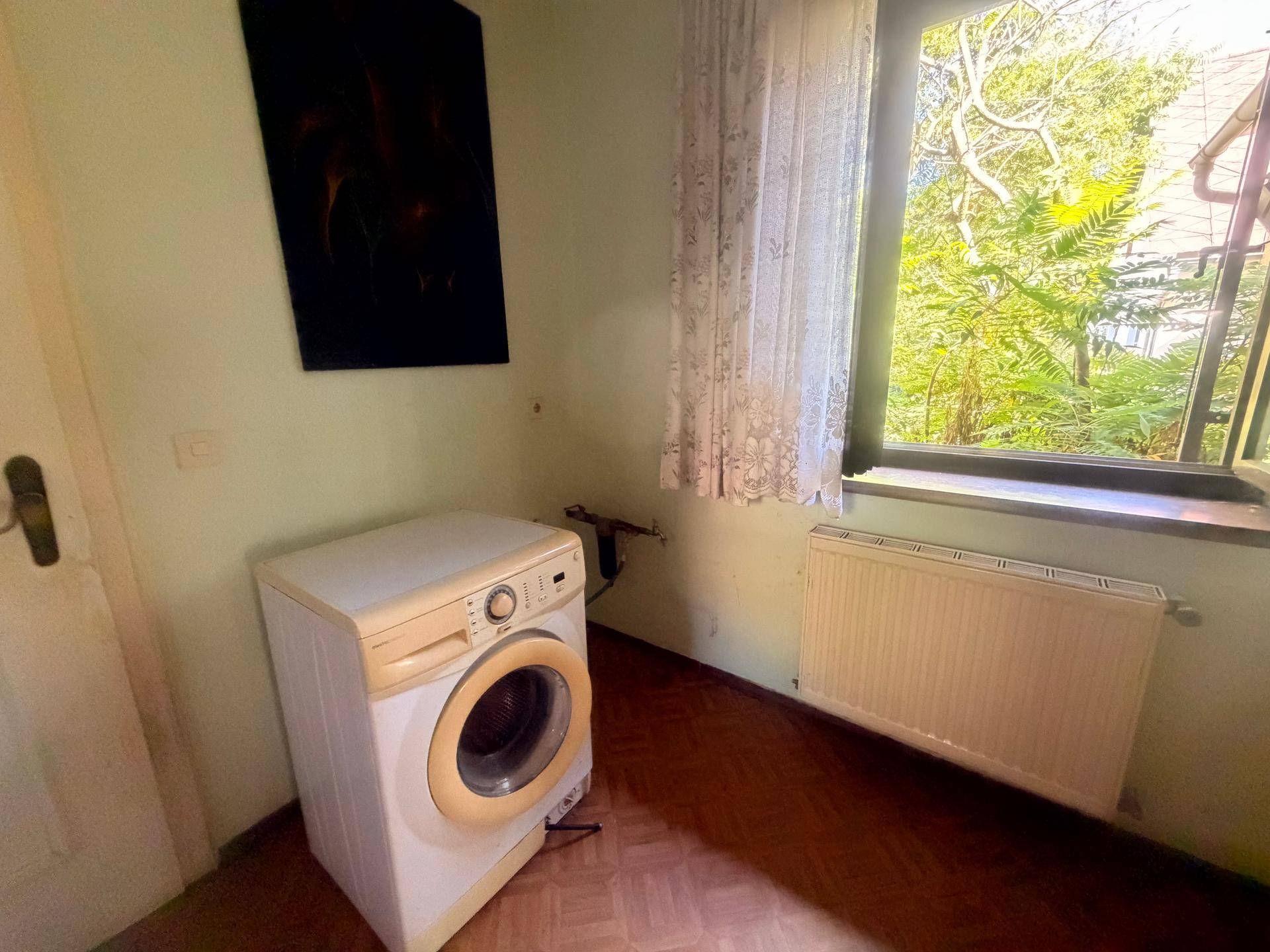 Kleiner Raum mit Waschmaschine und Heizkörper unter einem Fenster mit Vorhang.
