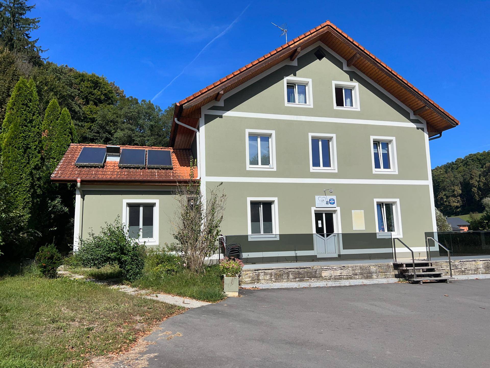 Gebäudeansicht mit Solarpaneelen auf dem Dach, großem Parkplatz und umgebender grüner Landschaft.