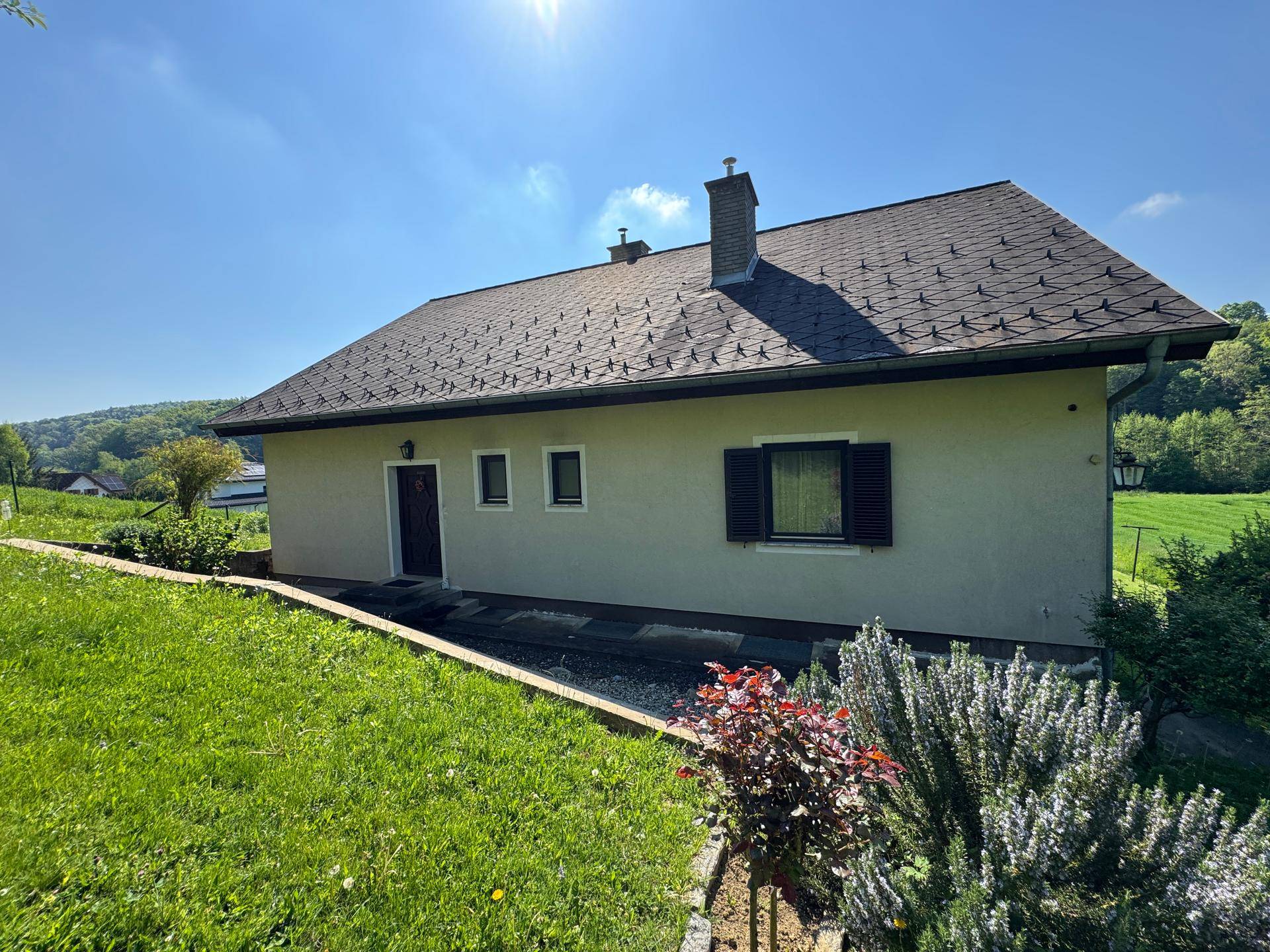 Seitenansicht eines Hauses mit Satteldach, grüner Rasenfläche und gepflegten Büschen.
