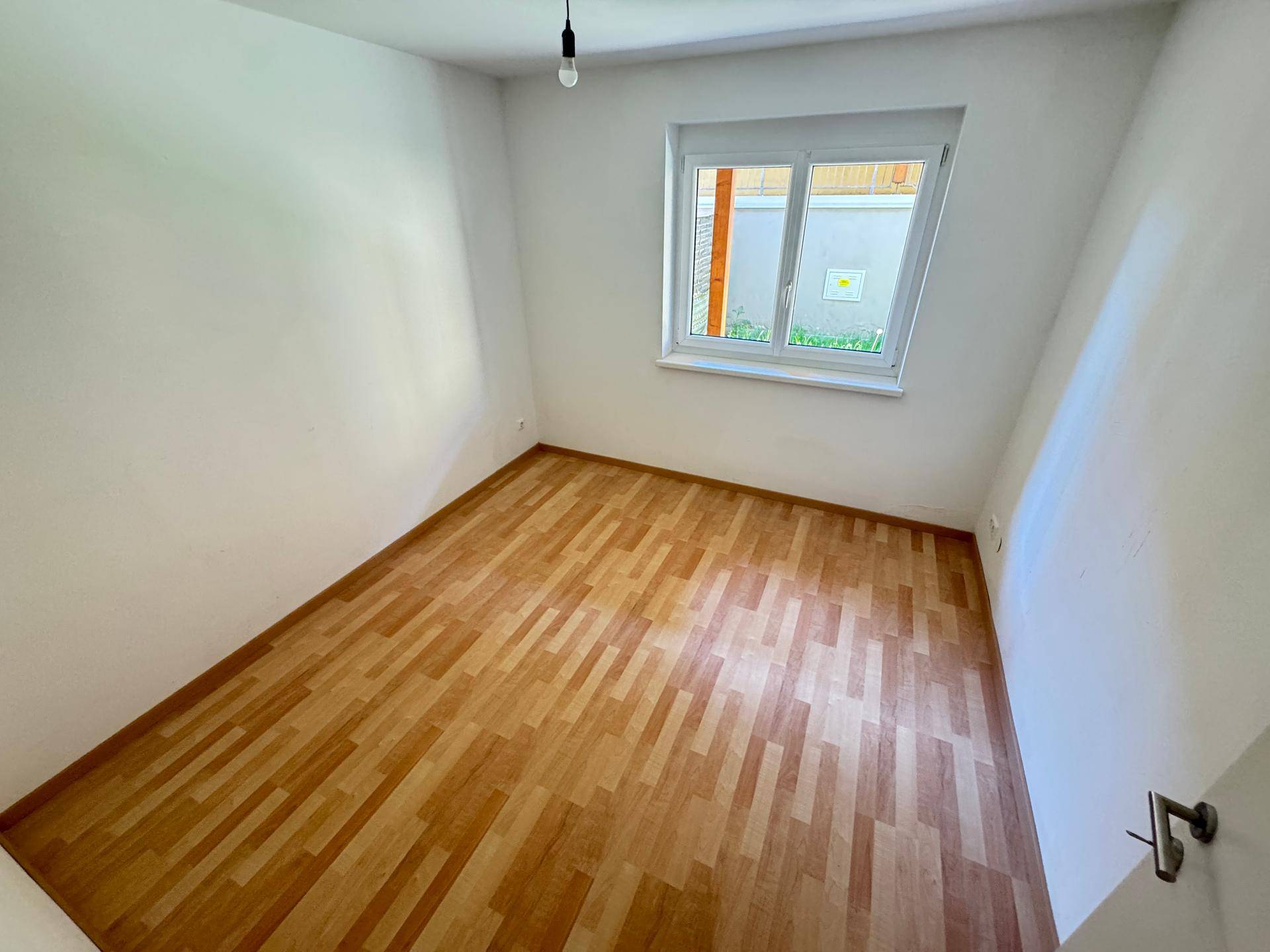 Leeres Zimmer mit hellem Laminatboden und einem Fenster, das viel Tageslicht hereinlässt.