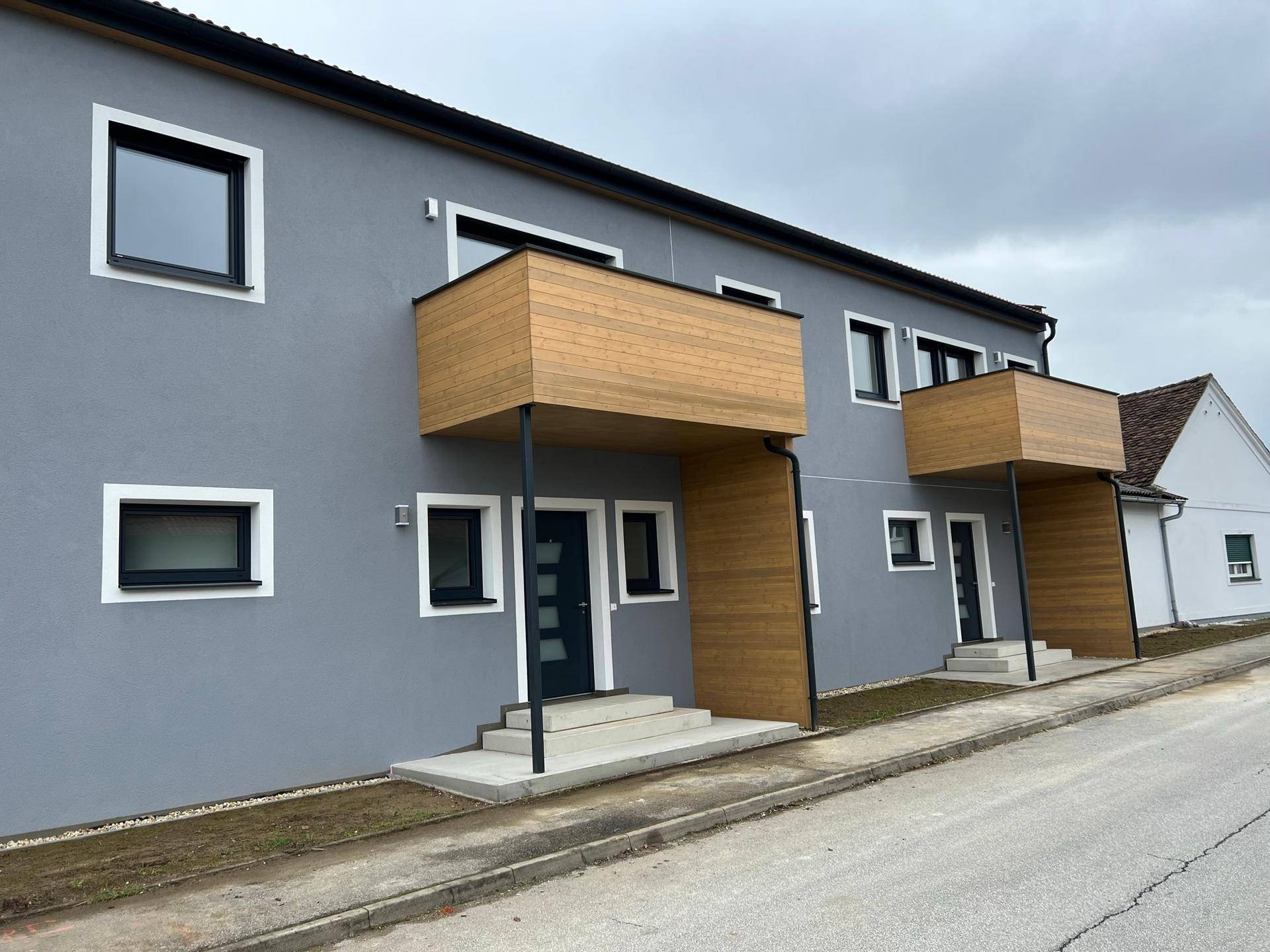 Modernes Wohngebäude mit grauer Fassade, Holzbalkonen und dunklen Fenstern in einer ruhigen Straße.