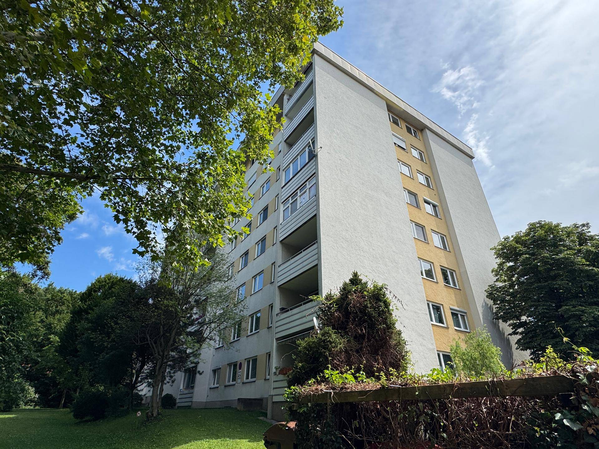 Mehrstöckiges Wohngebäude mit Balkonen, umgeben von Bäumen und Grünflächen unter blauem Himmel.