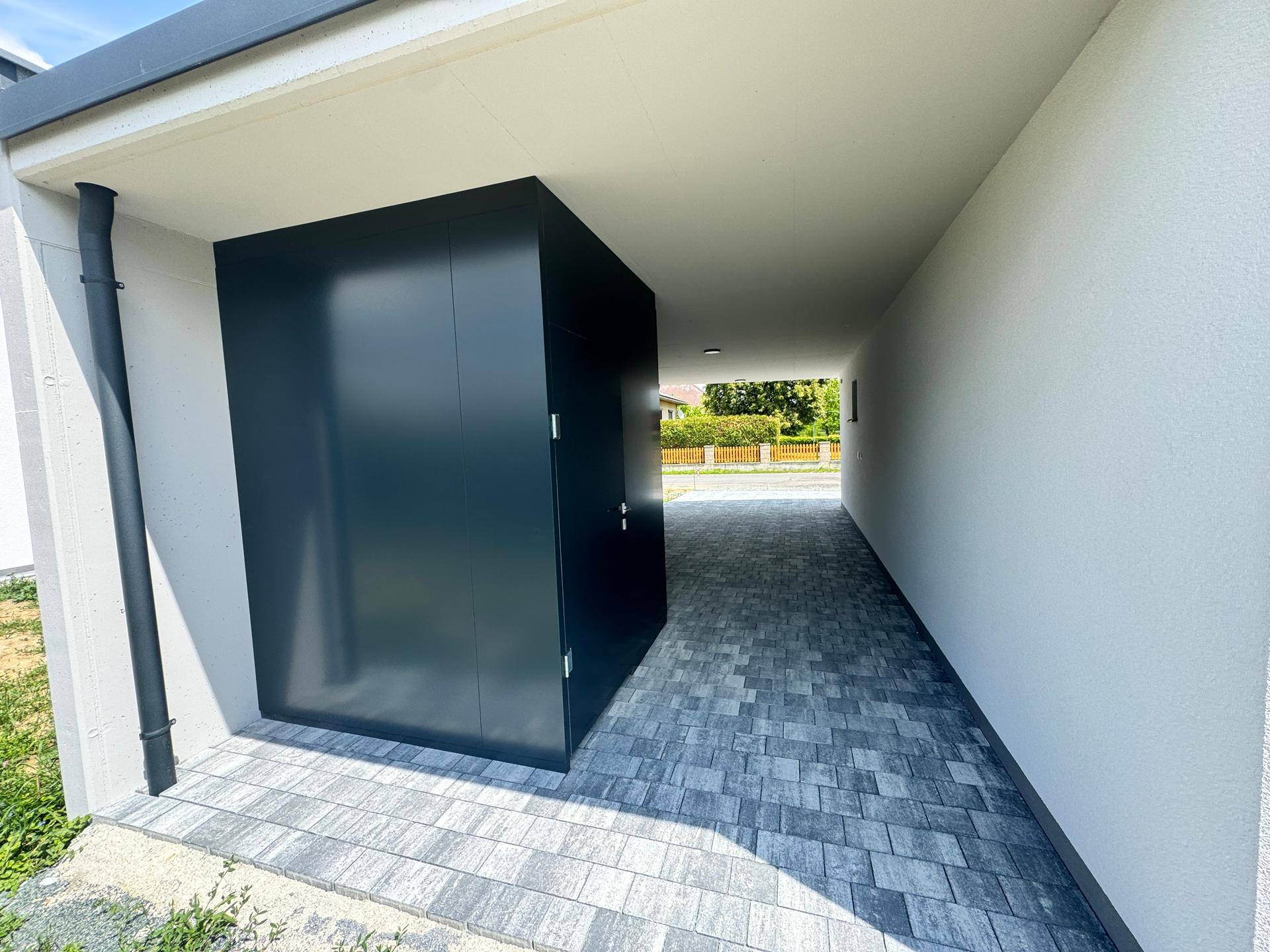 Carport mit integriertem dunklen Abstellraum oder Zugang, gepflasterter Bodenbelag.