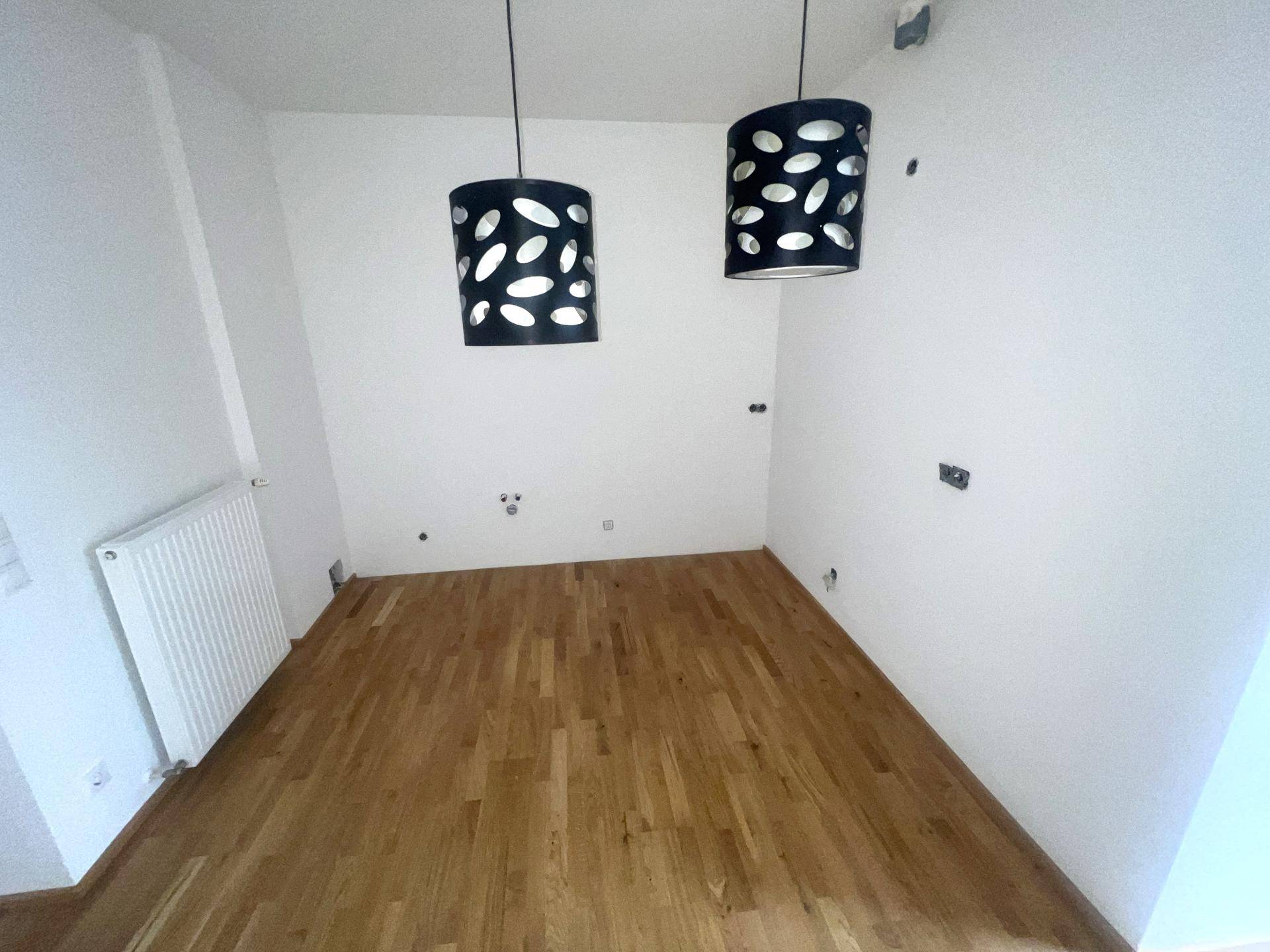Leerer Raum mit Parkettboden, Heizkörper und zwei modernen Deckenleuchten.