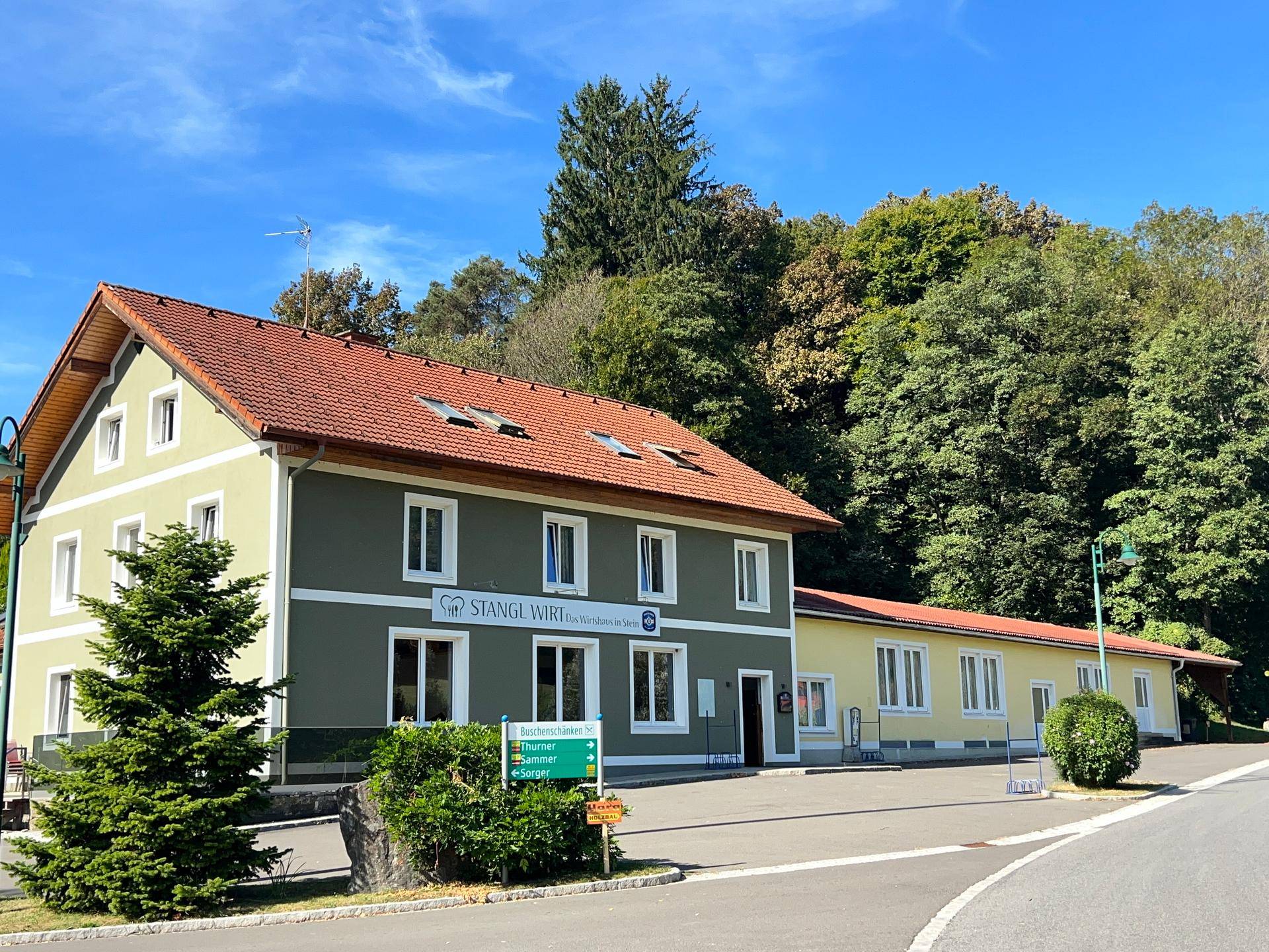 Traditionelles Gebäude mit grüner Fassade und rotem Ziegeldach, umgeben von Bäumen und einem Parkplatz.