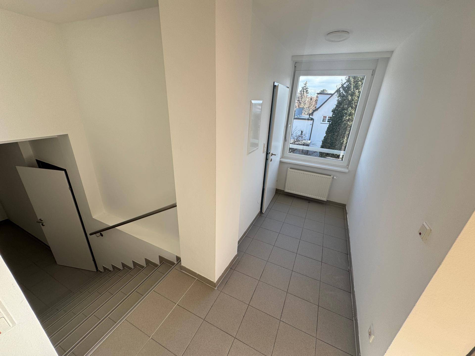 Heller Treppenaufgang mit Fliesenboden und Fenster mit Blick nach draußen.